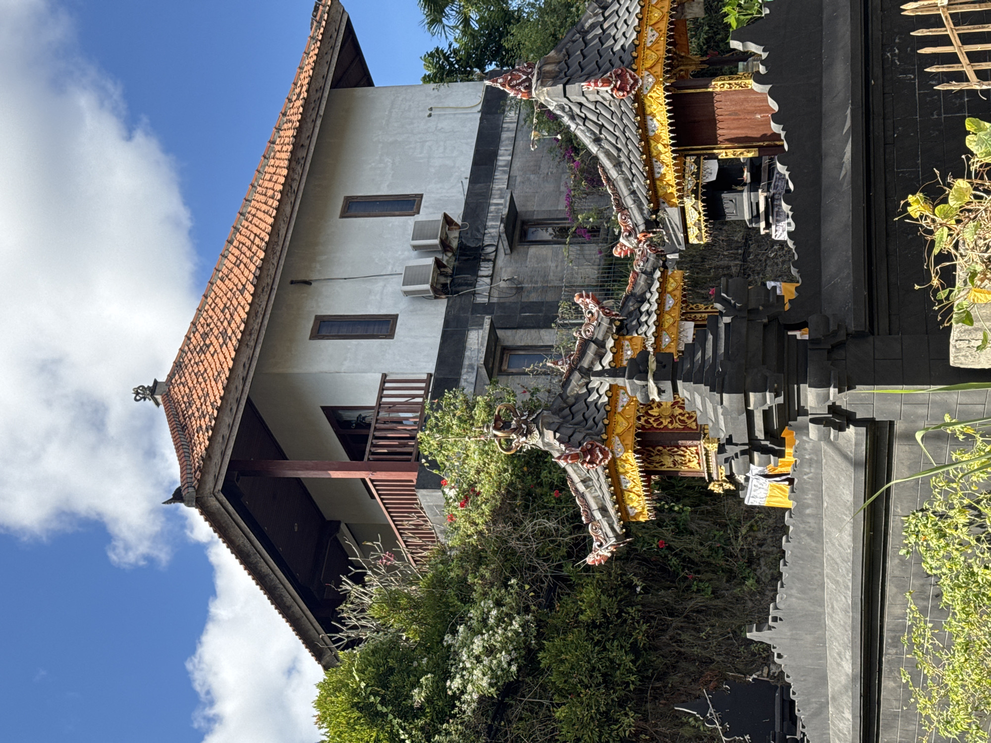 This is a medium shot photograph of a building and two small structures with ornate roofs, located in Kuta Selatan, Indonesia. The building in the background is a two-story structure with a white facade and a red tiled roof. It has a wooden balcony on the upper floor. There are two air conditioning units mounted on the exterior wall of the building. In front of the building, and partially obscuring the lower portion of the building, are two small, intricately decorated structures with dark tiled roofs and golden trim, characteristic of Balinese architecture. These structures are separated by a dark, textured stone wall. The area is surrounded by lush green vegetation, including trees and flowering bushes. The sky is a vibrant blue with scattered white clouds. The overall scene depicts a tropical setting with traditional Balinese architectural elements.