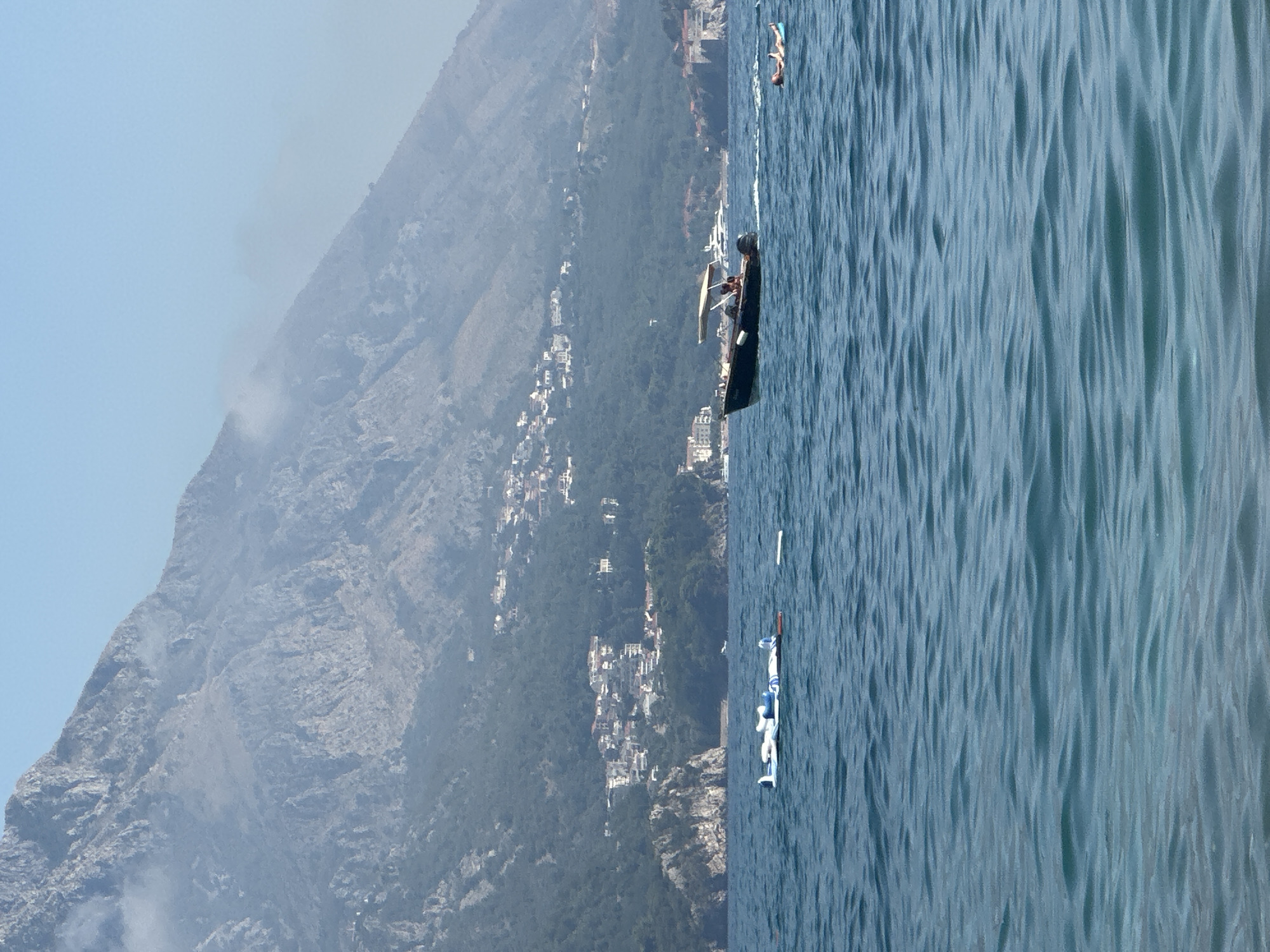 A wide shot captures the coastal scene of Boreti, Montenegro, on a hazy, bright day. The foreground is dominated by rippling blue water. In the middle ground, several inflatable objects, including what appears to be a deflated or partially deflated raft, float on the surface. To the right of these, a dark-colored motorboat with a canopy is underway, carrying at least two individuals. Further to the right, a person lies on a light blue inflatable mat in the water. In the background, a steep, verdant mountain rises, dotted with numerous white buildings forming a settlement. The mountain slopes are covered in dense green foliage, and patches of white haze are visible clinging to its upper reaches.