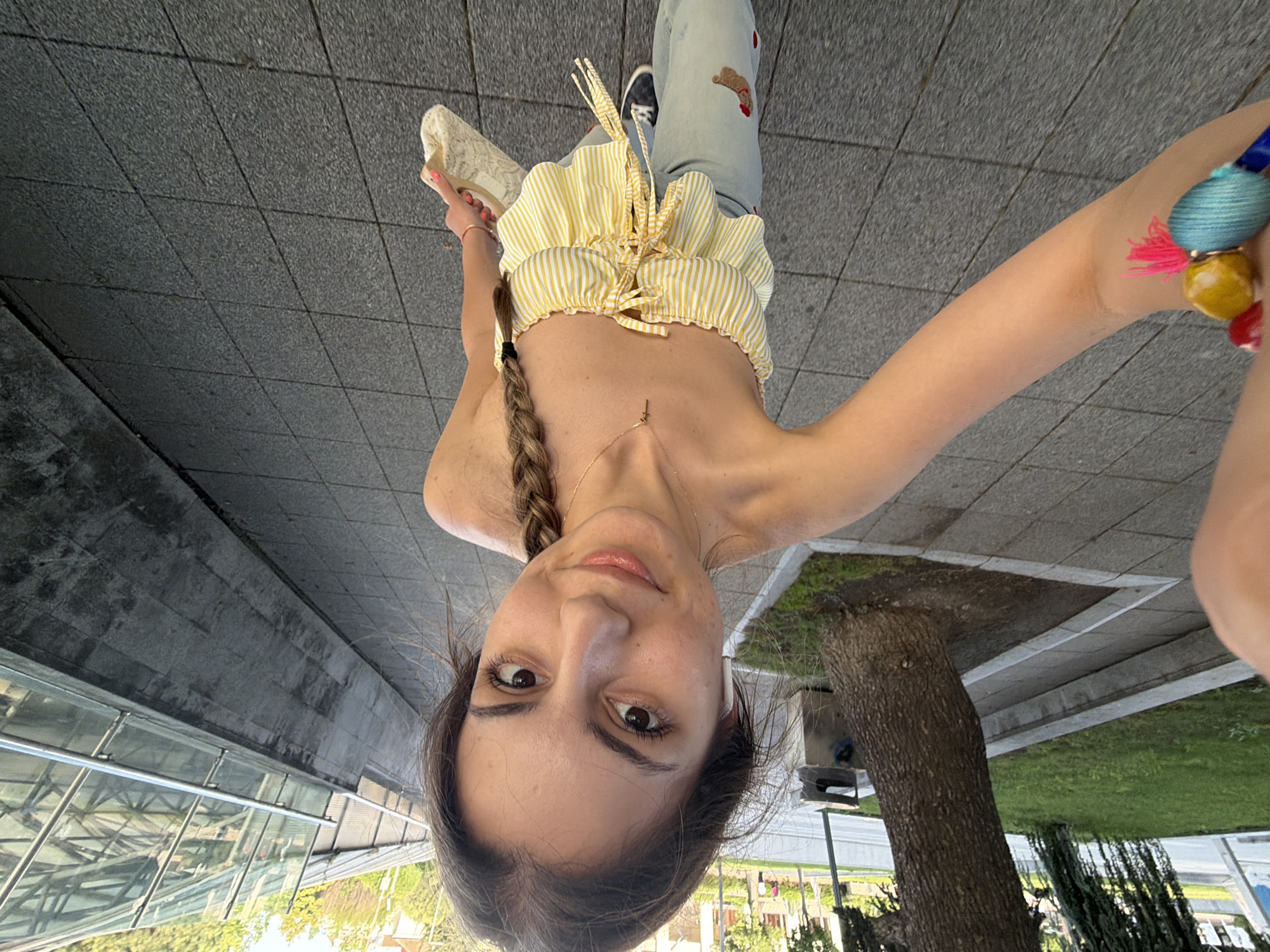 The image, originally oriented upside down, captures a young woman taking an overhead selfie in an outdoor urban setting in Burgas, Bulgaria. She looks directly at the camera with a neutral, slightly smiling expression. Her brown hair is styled in a braid over her left shoulder.
She wears a distinctive yellow and white striped ruffle crop top that exposes her midriff, and light blue jeans adorned with embroidered patches, including a red heart and a brown bear-like figure on the right thigh. A delicate gold necklace with a small charm is visible around her neck. On her left wrist, she wears a thin orange bracelet, while her right arm features multiple colorful, beaded bracelets with tassels in blue, yellow, red, and pink. A white earbud is visible in her right ear, and she is wearing black sneakers.
The ground beneath her is a dark gray paved surface, possibly concrete tiles. To her left (when viewed upright) is a modern building with large glass panels and metal railings, reflecting trees and sky. To her right, a tree with dark bark stands on a patch of green grass, bordered by more paving. The background reveals further greenery, distant buildings, and what appears to be a street or pathway, all illuminated by bright, clear daytime light. No visible text is present in the image.