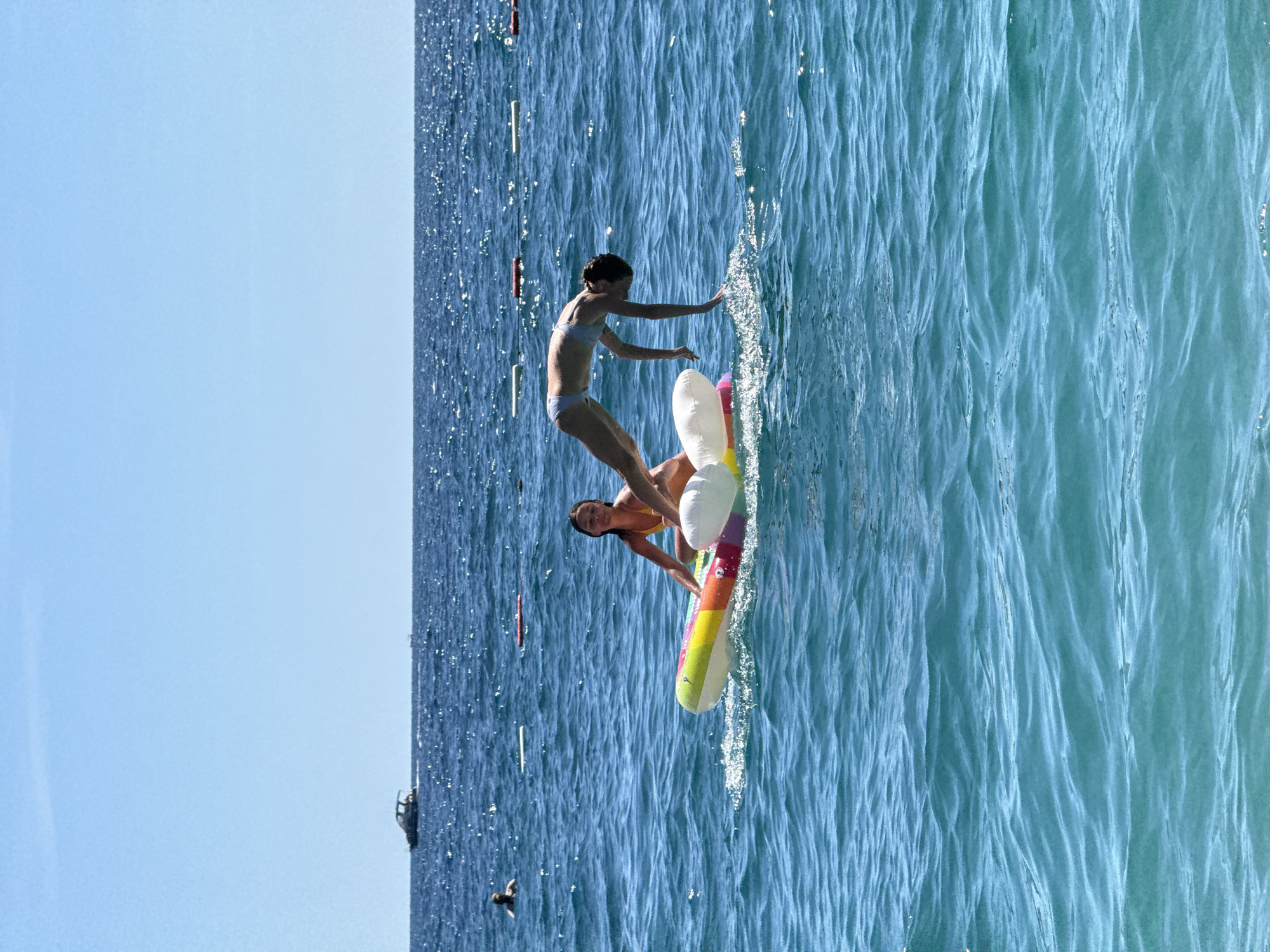 Two young women are enjoying a day at the beach in Kamenovo, Montenegro. The scene is set on a sunny day with clear blue skies and calm, turquoise water. One woman, wearing a light blue bikini, is standing on a colorful inflatable. She is leaning forward, with her arms extended for balance, as if she is about to dive into the water. Behind her, partially submerged, is another young woman in a yellow bikini, holding onto the inflatable. The water is a vibrant blue, with ripples and small waves creating a shimmering effect on the surface. In the distance, a boat is visible on the left side of the frame, suggesting it's a popular spot for water activities. The overall mood is one of leisure and fun, capturing a typical summer day at the beach. There is no visible text in the image.