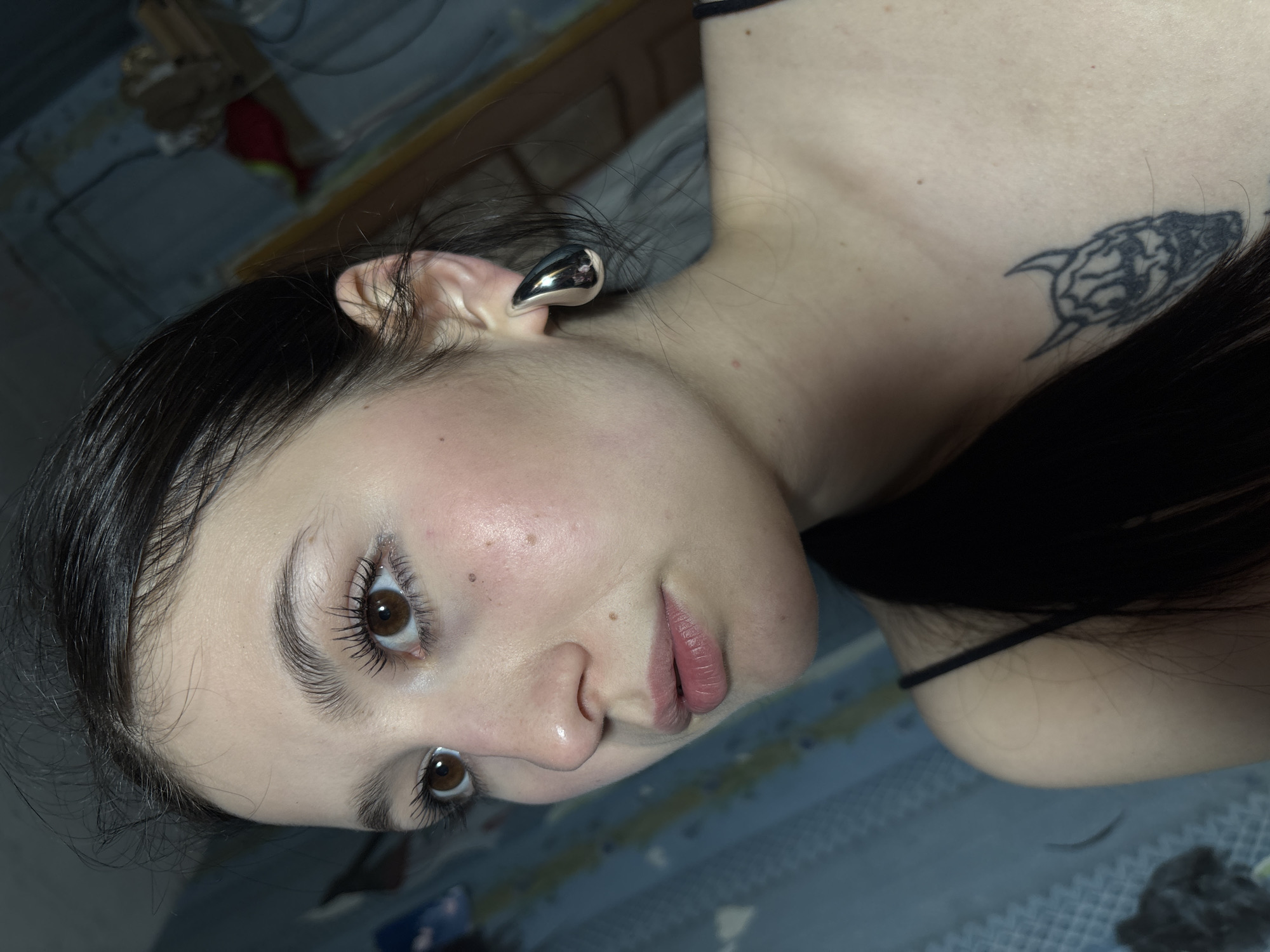 A close-up, rotated image of a woman with dark hair and brown eyes. She has fair skin with some freckles, and her lips are a light pink color. She is wearing a silver ear gauge in her left ear. A tattoo of a stylized animal (possibly a wolf or dog) is visible on her upper chest near her shoulder. The background appears to be a bedroom setting with a bed and some miscellaneous objects visible. The location in Фоки, Russia cannot be determined from the image.
