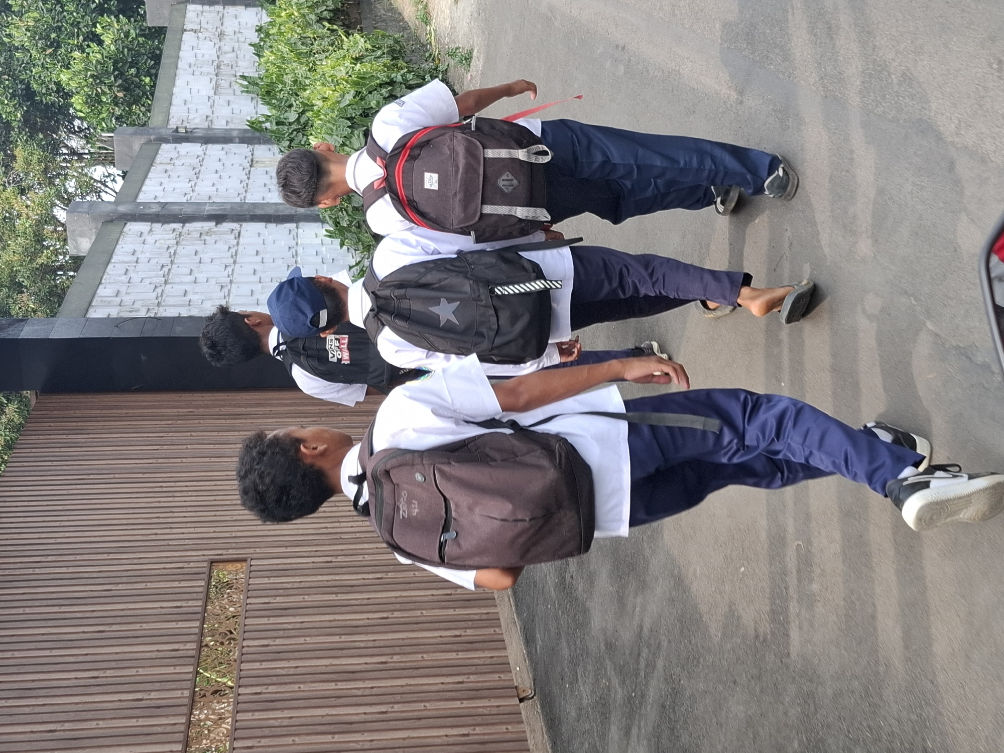 Four young males, likely students, are walking away from the camera on a paved outdoor path. The first student, on the left, wears a white short-sleeved shirt, blue pants, and black and white sneakers. He carries a dark brown backpack. To his right, a second student wears a white shirt, blue pants, and dark gray flip-flops, carrying a black backpack with a white star graphic. The third student, in the middle, wears a white shirt, blue pants, and dark gray flip-flops, carrying a black backpack with a blue baseball cap on top. The fourth student, on the far right, wears a white short-sleeved shirt, blue pants, and dark sneakers, carrying a dark brown backpack with red accents. They are walking alongside a tall, vertical wooden fence on the left and a light-colored textured wall with greenery on the right. The environment suggests a sunny day in Balekambang, Indonesia, possibly near a school.
