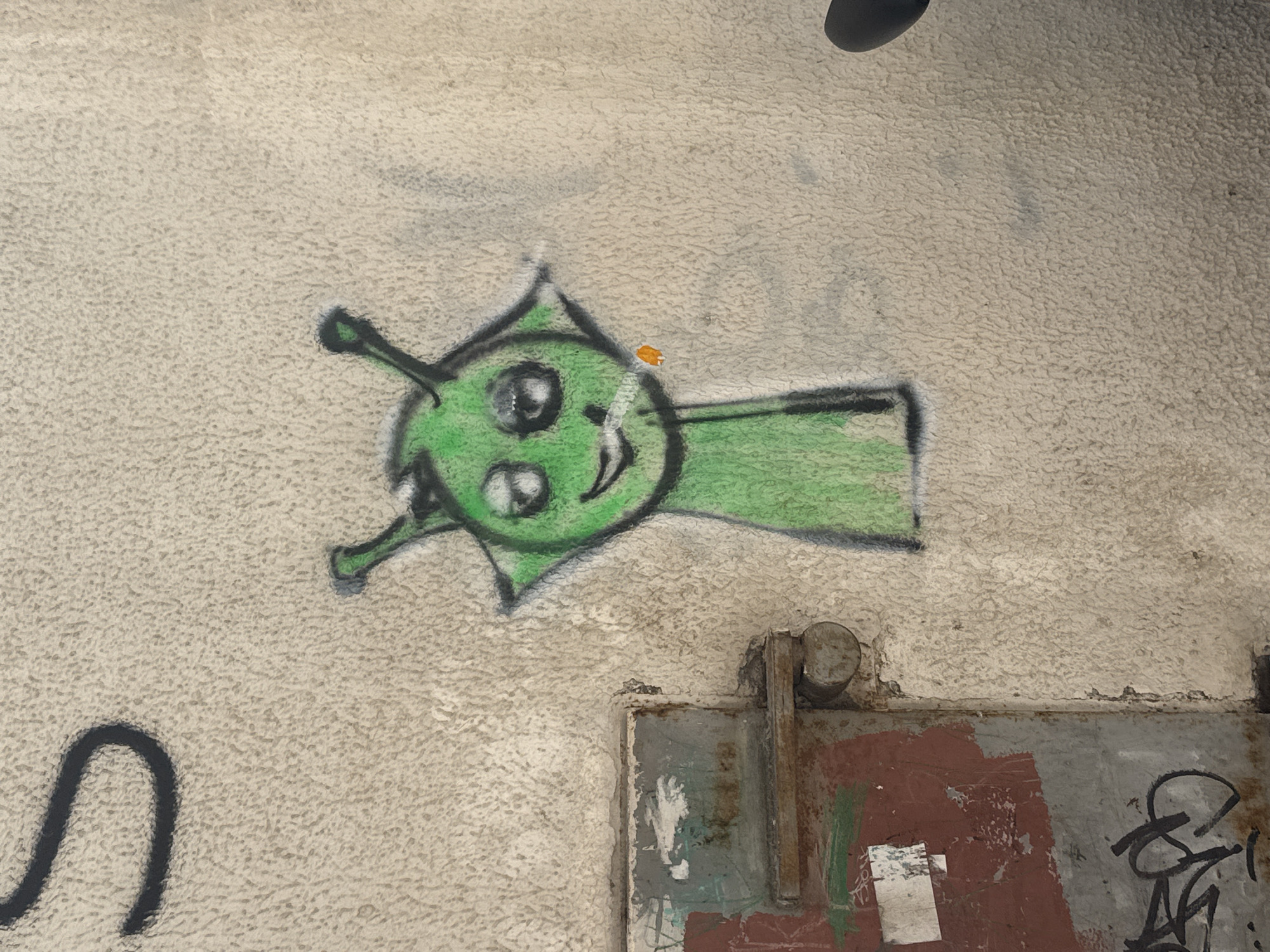 A green, alien-like cartoon figure is spray-painted on a textured, light-colored wall. The figure has a triangular head with two eyes, a smiling mouth, and antenna-like protrusions on either side. A horizontal green shape extends from the right side of the head, resembling a tail or appendage. The cartoon is outlined in black.
Below the figure is a partially visible metal door frame, colored with a mix of grey, red, and brown paint. Some graffiti is present on the metal surface. The wall's texture is rough and uneven.
Based on the prompt, the location is in the city of Šušanj, Montenegro.