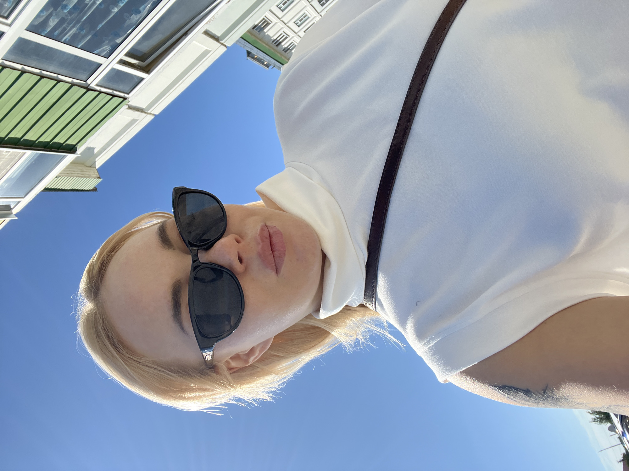 The image is a close-up, low-angle selfie taken outdoors on a sunny day. The primary subject is a woman with blonde hair wearing black sunglasses and a white shirt. She is looking up towards the camera, and her lips are slightly parted. A thin, dark strap, likely from a bag or garment, is visible across her shoulder. The background consists of a clear, bright blue sky and the upper portion of a building with multiple windows and green siding. The lighting suggests it is daytime, possibly late morning or early afternoon. The overall mood appears casual and relaxed. There is no visible text in the image.