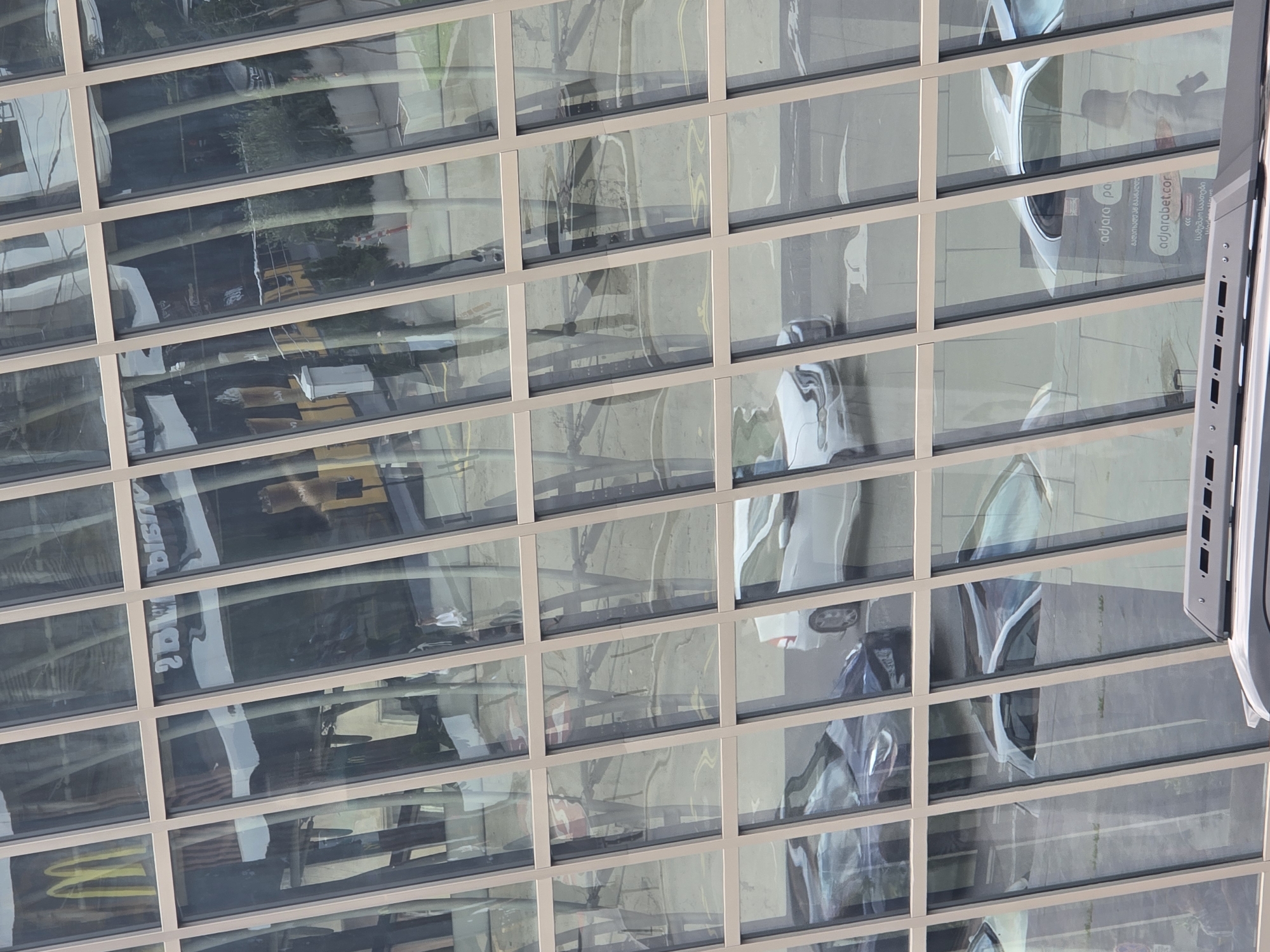 The image displays a reflection on the facade of a modern building in Tbilisi, Georgia. The building's exterior is composed of large, angled glass panels framed by a light-colored structural material. The reflections show a street scene with cars, including white sedans and a darker vehicle. A McDonald's sign with its characteristic yellow arches is visible in one of the reflections, suggesting proximity to a restaurant. A portion of a website, "adjarabet.com", is also reflected on the glass. The scene is captured from an elevated perspective, looking down at the street and its reflected elements. The lighting suggests it is daytime, with sunlight creating highlights and shadows on the reflective surfaces.
