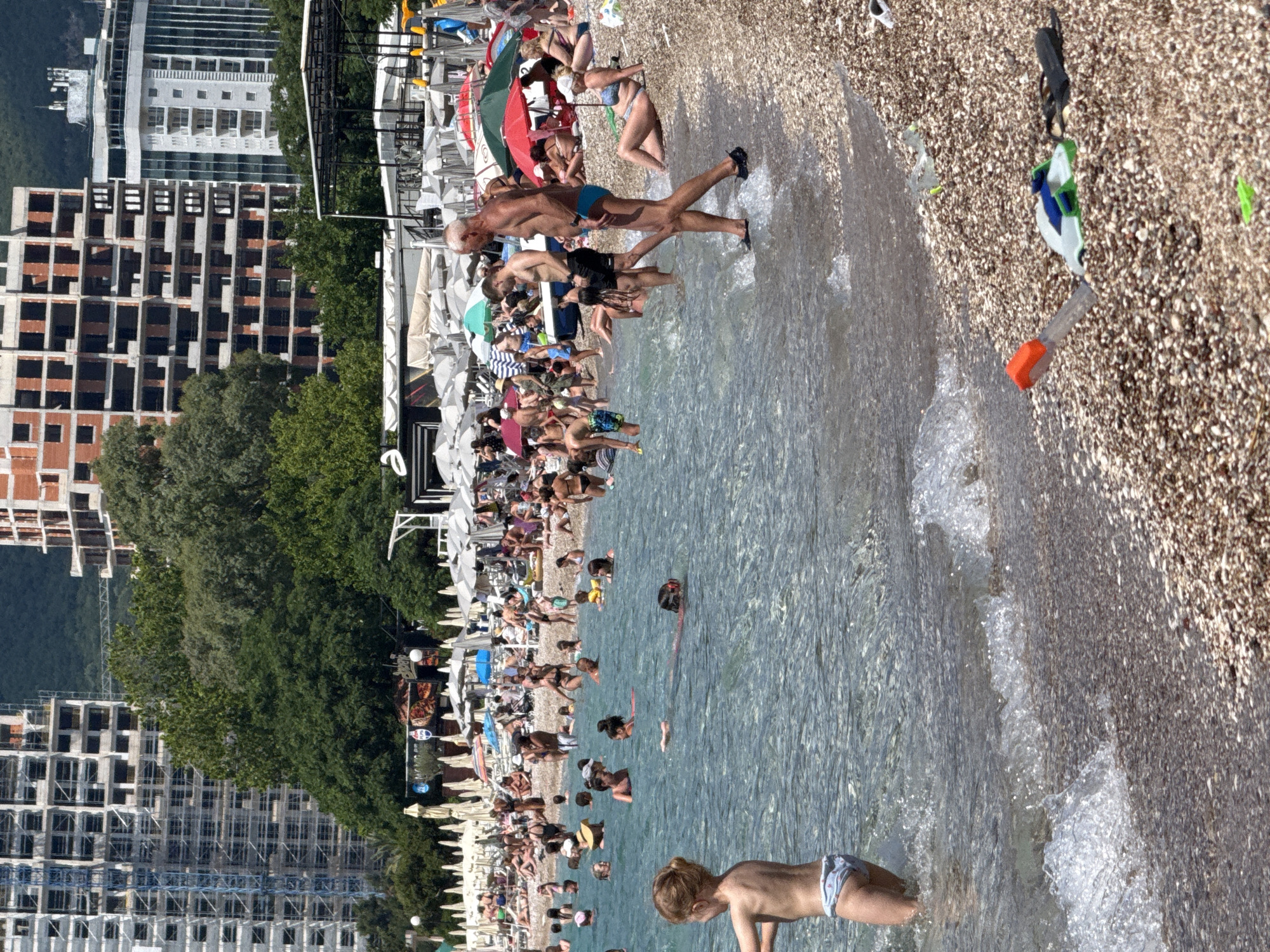 The image depicts a crowded beach scene in Bečići, Montenegro, with numerous people enjoying the water and the shore. In the foreground, a young girl with blonde hair is standing in shallow water, facing away from the camera. To her left, further out in the water, a person wearing a mask and snorkel is swimming. The beach is covered in pebbles and populated with many sunbathers and swimmers. Several beach umbrellas are set up, providing shade for some of the beachgoers. In the background, large, unfinished buildings and trees are visible, suggesting ongoing construction in the area. The water is clear and a bright blue. Several objects are scattered on the pebble beach, including a snorkel and mask set.