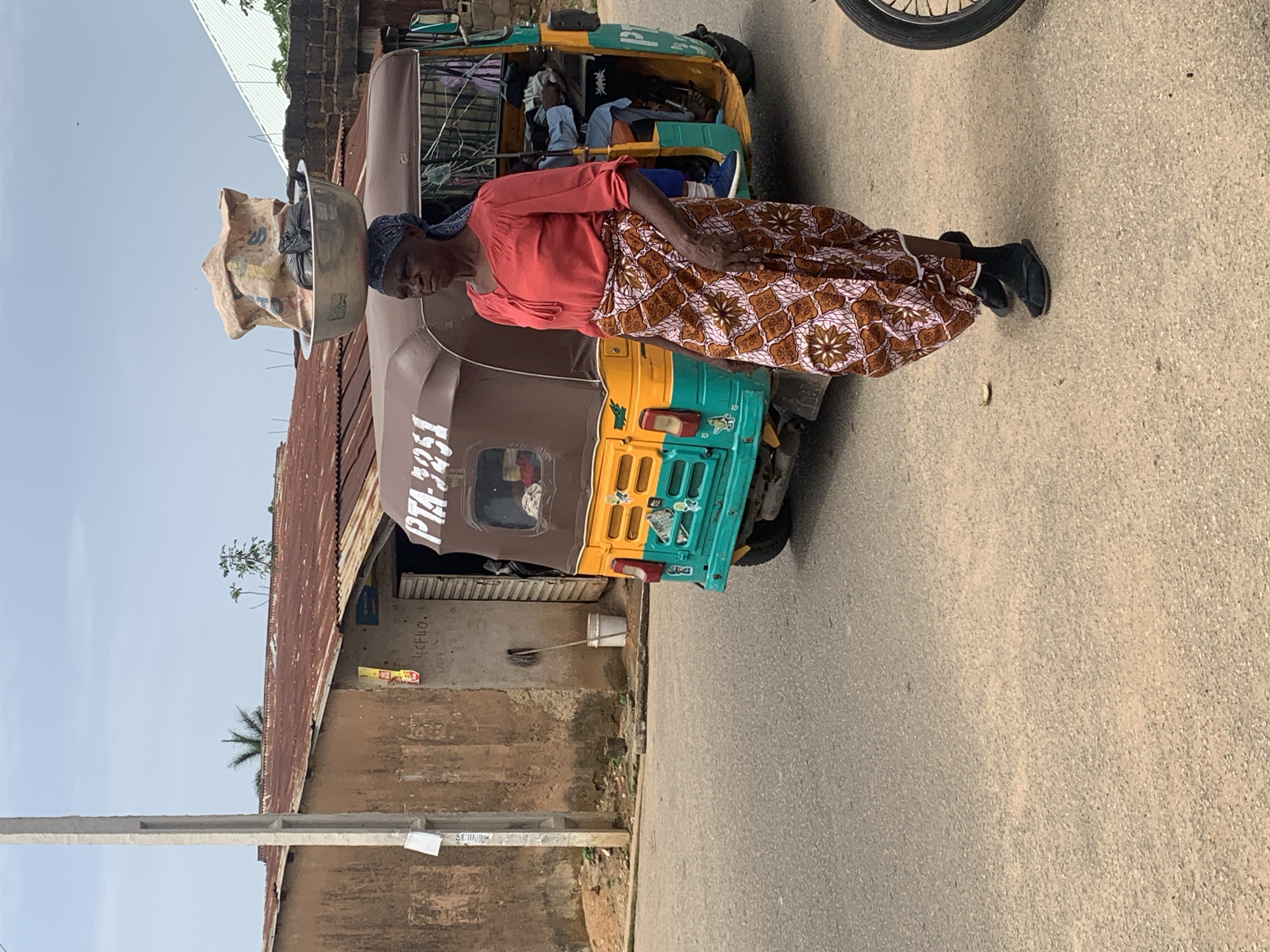 A woman, likely of African descent, is depicted with her head bowed, carrying a large bowl filled with goods, possibly produce or grains, on her head. She is wearing a coral-colored, long-sleeved blouse and a patterned skirt in shades of brown and cream. Her attire suggests a traditional or casual style. She appears to be standing next to or in front of a three-wheeled vehicle, commonly known as a "keke" or "marwa" in Nigeria, which is partially visible. The vehicle is yellow and green with some brown sections. Inside the keke, other people are seated, their faces obscured by distance and the angle of the shot. The scene takes place on a paved road, with a weathered, corrugated iron roofed building on the left side of the image. The building has a rustic appearance, with peeling paint and visible structural elements. The sky is overcast, suggesting it might be daytime, but the quality of light is not strong. The overall setting appears to be a street scene in a populated area, possibly a market or a transportation hub, typical of urban or semi-urban Nigeria. The woman's posture and the load on her head suggest she is engaged in a common activity of transporting goods for sale or personal use. There is some visible text on the side of the keke, appearing to read "PTA-3251." Another smaller text appears on the building wall. The image is oriented vertically, with the top of the frame pointing to the right.
