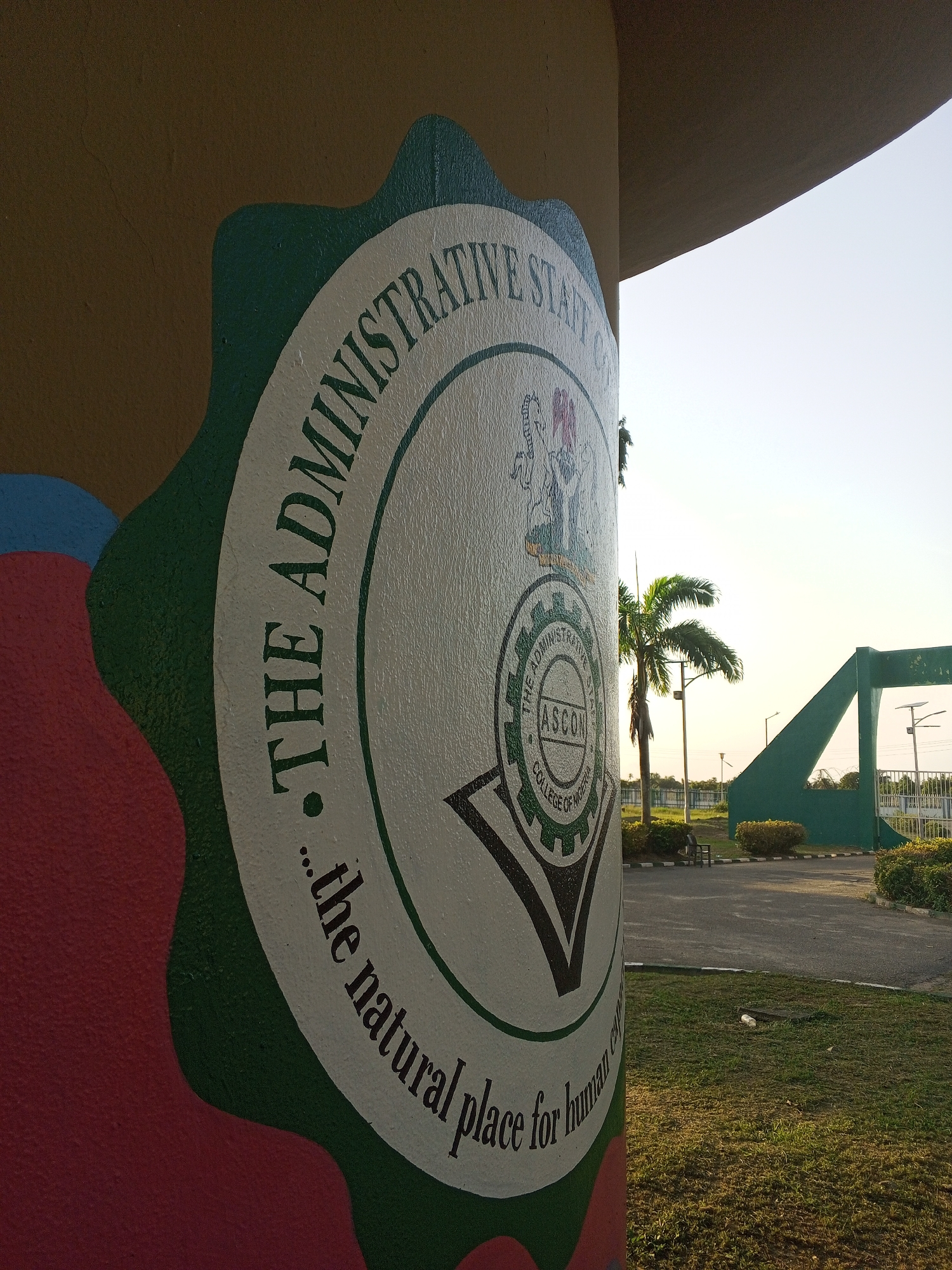 The image shows the logo of the Administrative Staff College of Nigeria (ASCON) on a pillar in the city of Tawpo, Nigeria. The logo is a circular design with the college's name surrounding a central emblem containing the letters ASCON and the words "College of Nigeria". The Nigerian coat of arms is positioned above the central emblem. The pillar is decorated with colorful paint around the logo. In the background, the college's entrance gate, palm trees, and other landscaping are visible under a bright sky.