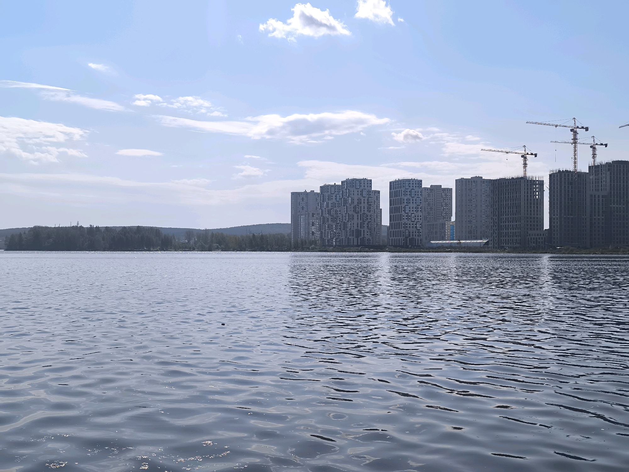 The image depicts a wide, calm body of water in the foreground, with ripples reflecting the light. In the background, a line of trees borders the far side of the water, beyond which a cityscape is visible. The city skyline is dominated by numerous tall, modern residential buildings under construction, many of which are incomplete, with visible scaffolding and cranes. Three large tower cranes are prominently positioned among the buildings, indicating active construction work. The sky is partly cloudy with a pale blue hue. The scene is likely located in Yekaterinburg, Russia, given the architectural style of the buildings and the presence of construction cranes, indicative of the rapid development often seen in Russian cities.