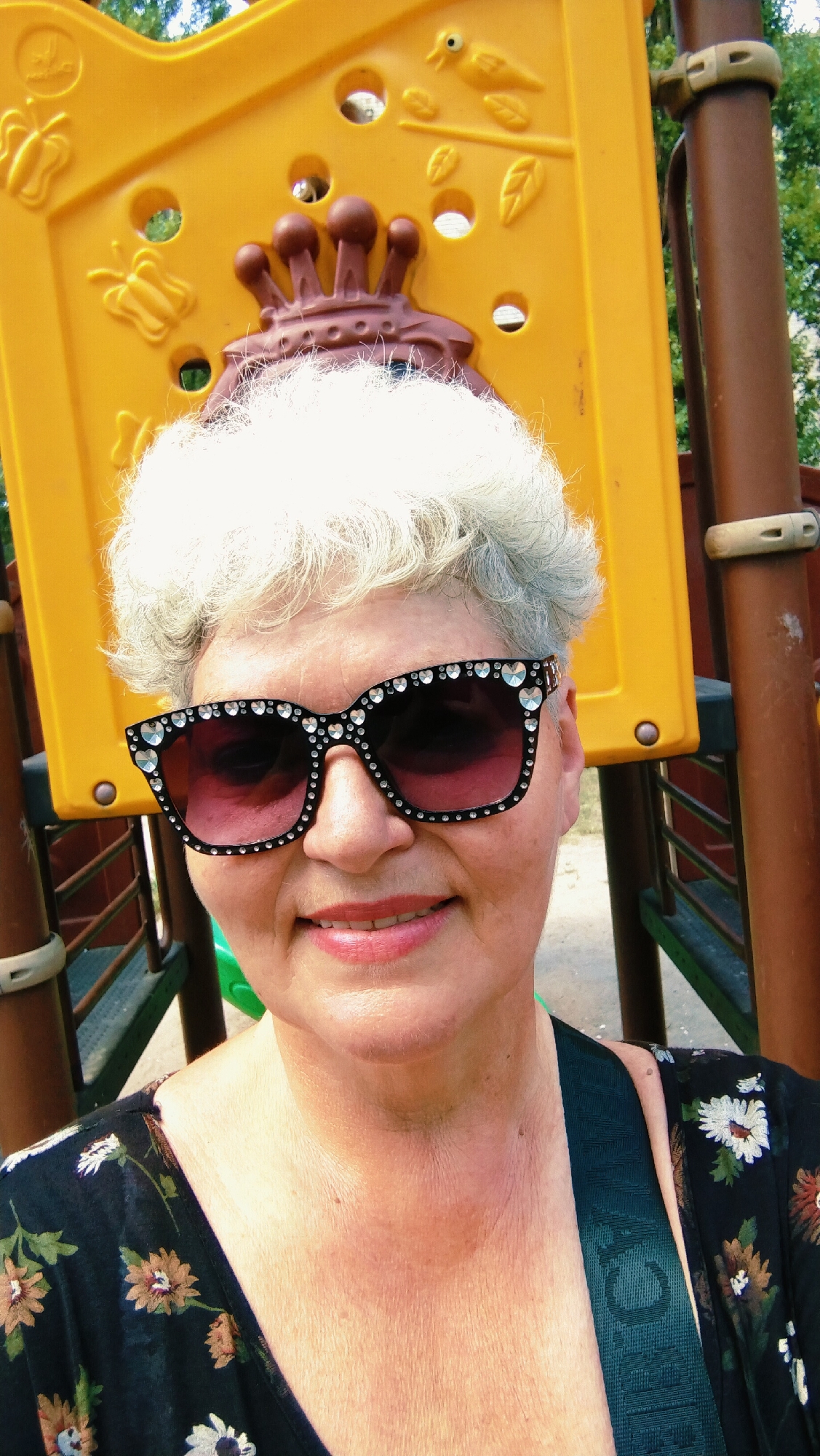 The image shows a close-up, selfie-style portrait of a woman with short, white, curly hair and fair skin. She is wearing large, black, cat-eye sunglasses adorned with rhinestones. Her lips are painted a glossy pink. She is smiling. The woman is wearing a black top with a floral pattern featuring white and orange flowers. Behind her, a yellow playground structure with brown accents is visible. The environment appears to be outdoors, possibly a park or a backyard, with hints of greenery and trees in the background. The scene is set in the city of Horishni Plavni, Ukraine.