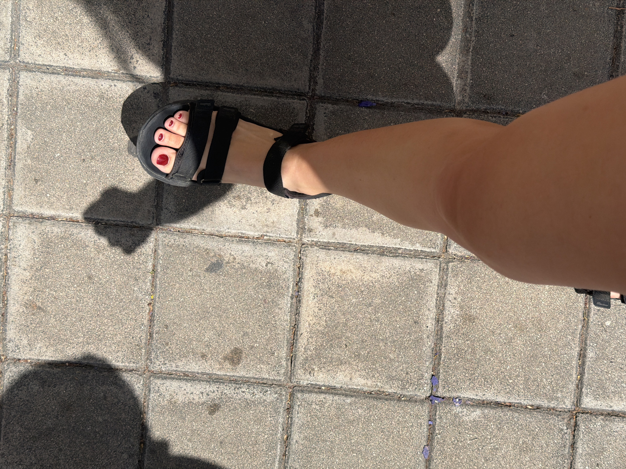 A woman's right leg and foot are shown, wearing a black sandal with red nail polish on her toes. The foot is positioned on a paved walkway made of square grey tiles. Shadows from an overhead structure are cast across the walkway. The scene is in Alicante, Spain.
