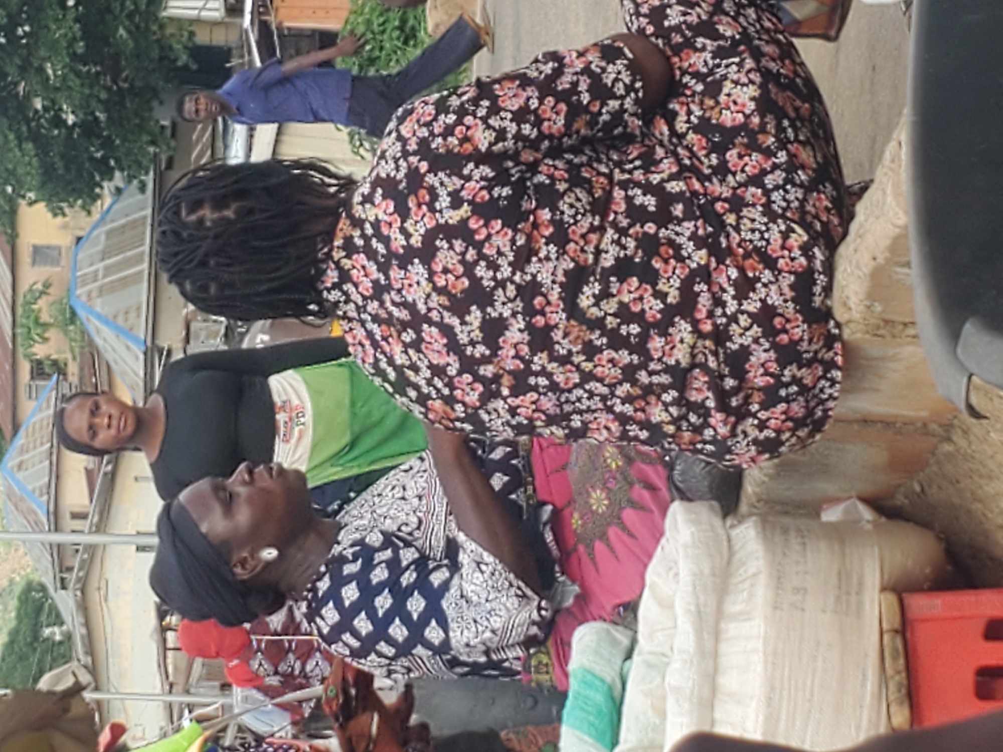 In Gwong, Nigeria, a street scene unfolds with several individuals. In the foreground, a woman wearing a black headscarf and a patterned blue and white top sits, facing left. She has large white earrings. To her right, partially obscuring her, is another person from behind, wearing a dark top with a floral print. To the left of the seated woman, another person in a red headscarf is visible. Further back, a woman in a black top stands, and next to her, a green flag with "PDP" printed on it is displayed. In the background, a young man in a blue shirt and dark trousers walks away from the camera. The environment appears to be a market or roadside area with buildings and trees visible.