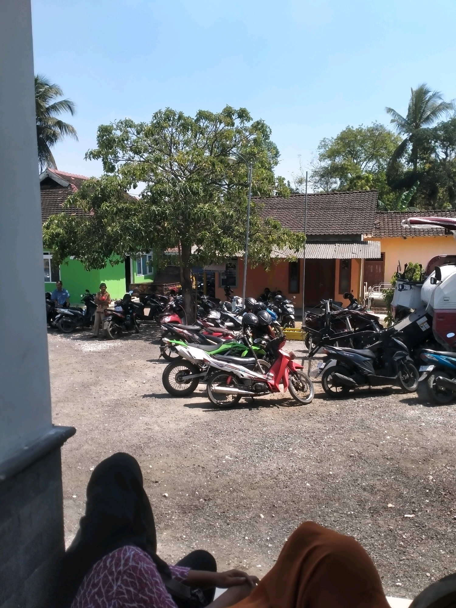 The image depicts an outdoor scene in what appears to be a parking lot in Playen, Indonesia. The foreground features the legs and lower torsos of two individuals seated, with one wearing a black headscarf and a patterned purple garment, and the other wearing an orange head covering and a brown garment. A shadow is cast on the ground from a structure on the left. The mid-ground is dominated by a large number of parked motorcycles of various colors including red, green, white, and black. A few people are visible in the distance near a green building, with two men standing. One man is wearing a blue shirt, and the other is wearing a striped shirt and is holding something. Behind the motorcycles, a row of buildings with tiled roofs is present. Trees, including a large one with dense foliage providing shade over some of the motorcycles, are scattered throughout the scene. The sky is clear and blue, suggesting a sunny day. The ground surface is covered in gravel. In the background, more trees and parts of other buildings are visible, including a white vehicle with red accents resembling a delivery truck or mobile vendor.