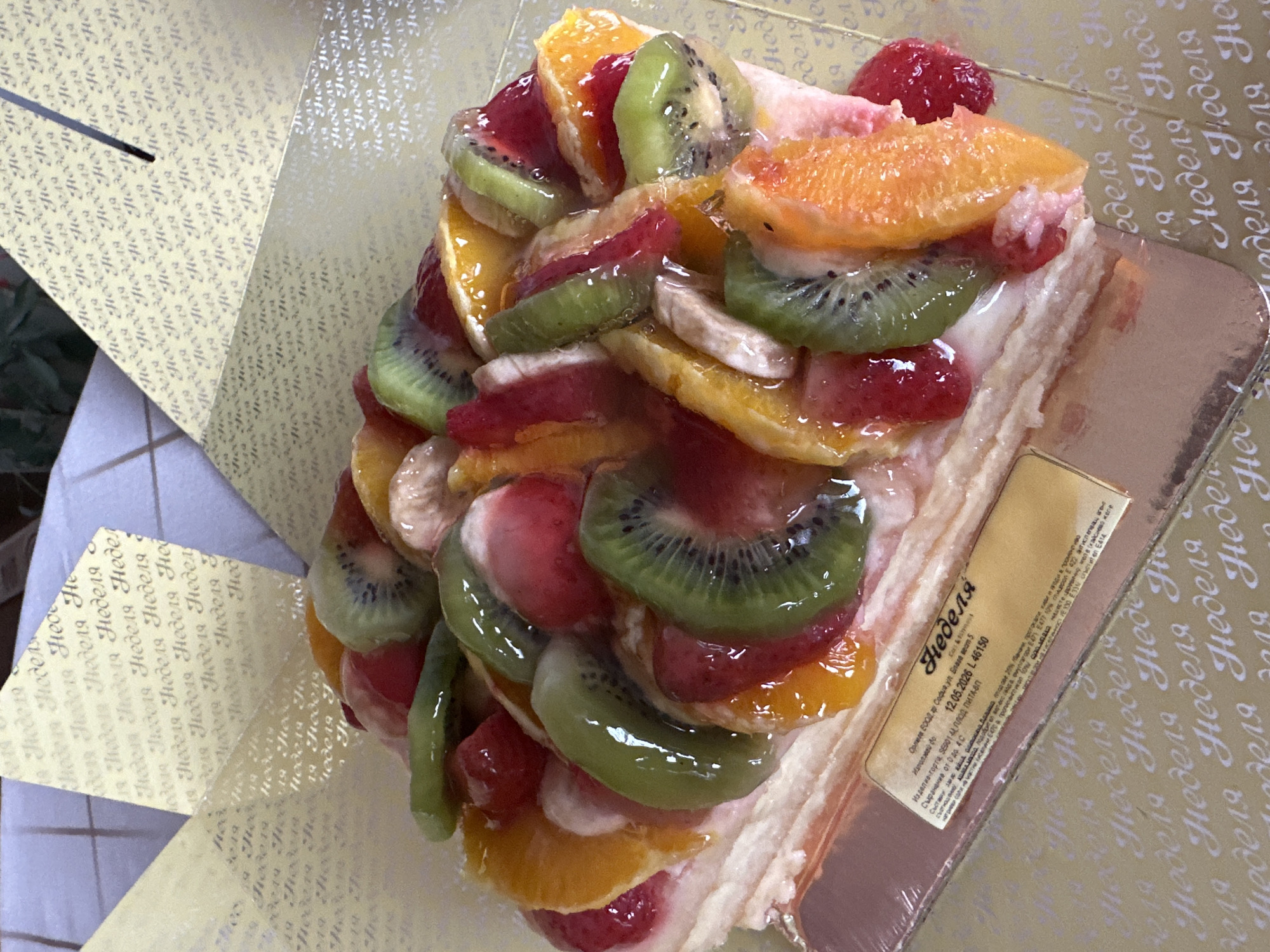 A piece of fruit-topped cake sits on a cardboard base with a label and is surrounded by a patterned paper box. The cake has layers of cream and is topped with sliced kiwi, strawberries, oranges, and bananas, coated with a clear glaze. The label has text in Cyrillic, which includes "Неделя" (Nedelya) and "Орлиев ЕООД, гр. София" (Orliev EOOD, Sofia). The patterned box has repeated Cyrillic text. The cake rests on a surface with a white and green speckled pattern. There is no indication of the location being in Montana, Bulgaria.