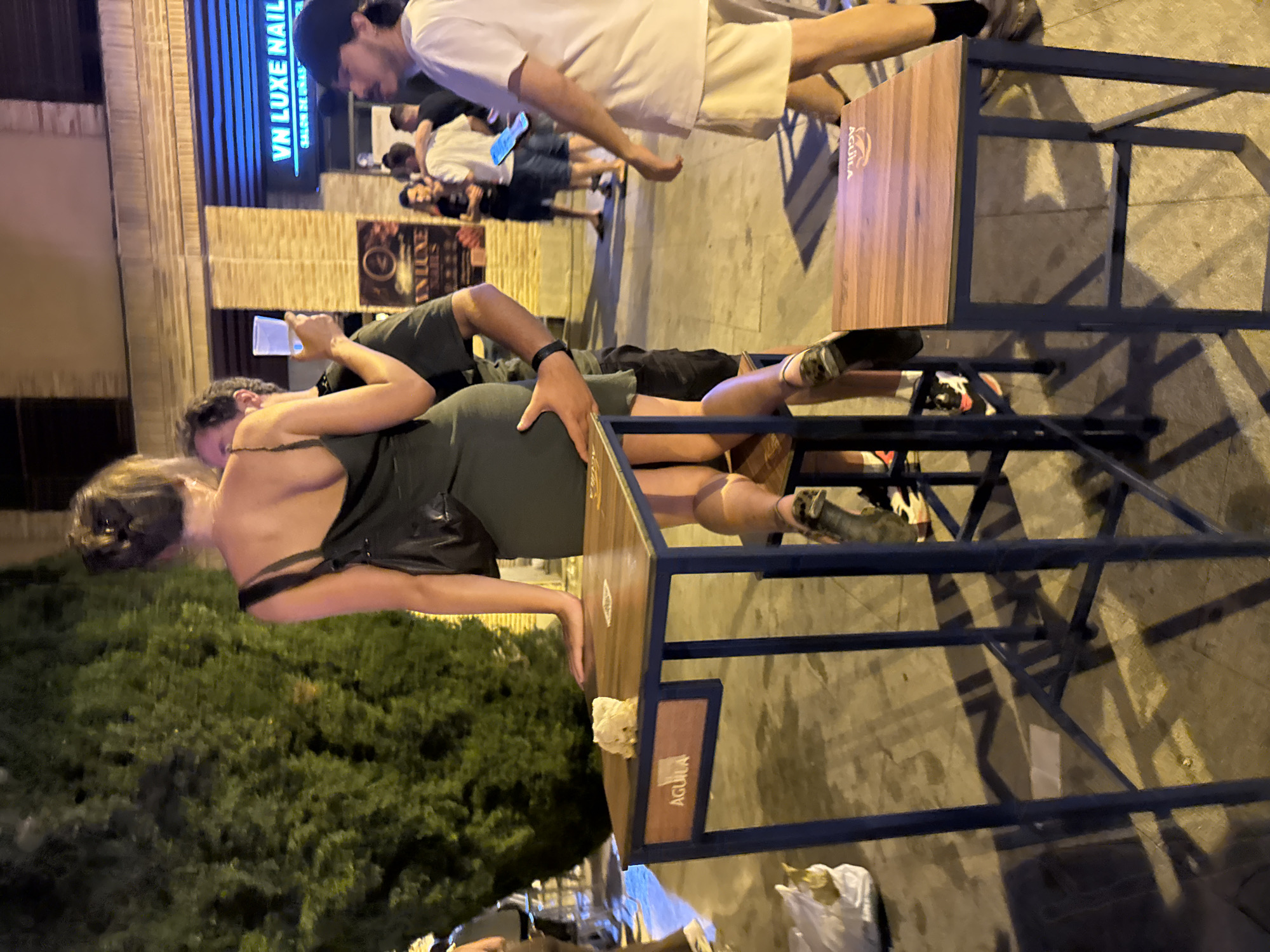 The image captures a street scene at night in Las Rozas de Madrid, Spain, near the "VN LUXE NAILS" establishment. In the foreground, a woman in a dark green dress is leaning against a tall, blue-legged table with a wooden top. Her right hand rests on the table, and a man in a dark t-shirt has his left arm around her waist. The woman is holding a clear cup with a pale liquid. To her left, a white bag lies on the ground. Another tall table and a stool are visible to the right, both bearing the "AGUILA" logo. In the background, other people are standing and walking on the sidewalk. A large, green tree is to the left of the frame. The lighting suggests artificial illumination from streetlights and the businesses.