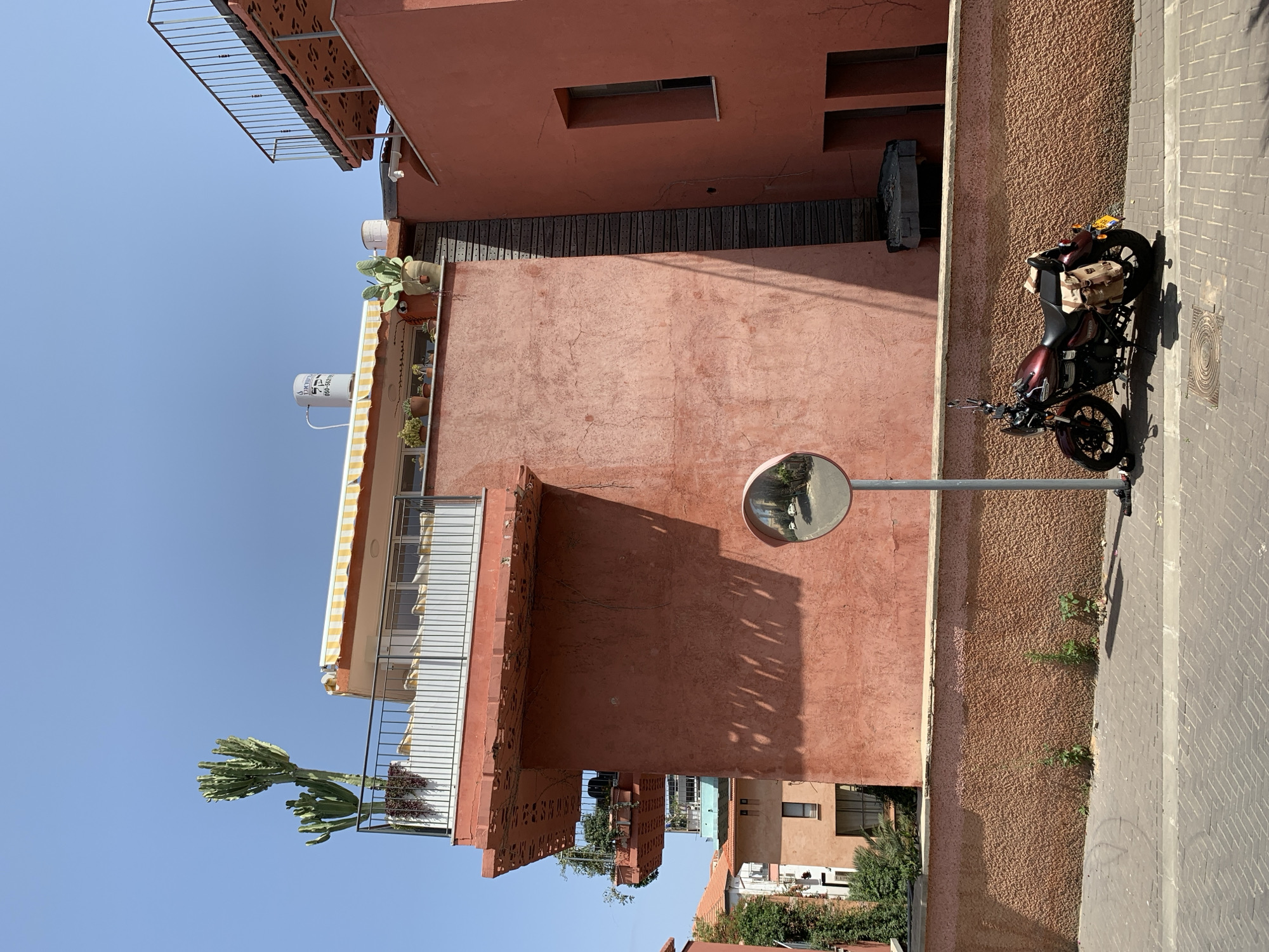 The image depicts a street scene in Tel Aviv, Israel. The architecture features buildings with reddish-brown exteriors and balconies. A motorcycle is parked on the street next to a textured wall. A round safety mirror is visible on a pole. The sky is clear and blue. Various plants and cacti adorn balconies and rooftops.