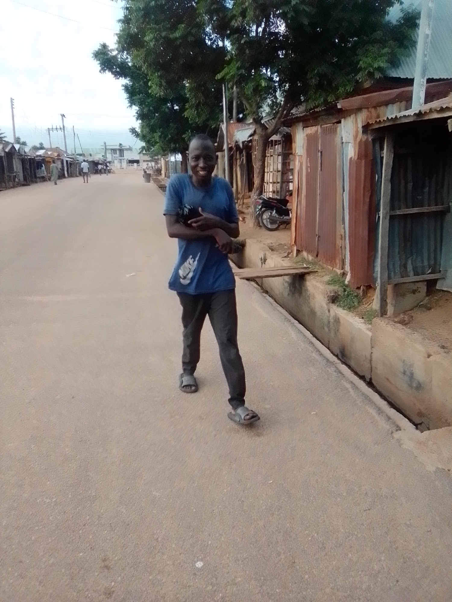 A full shot captures a young male standing on a dirt road in Gombe, Nigeria. He is wearing a blue t-shirt with a white Nike logo and dark pants. The man is smiling, holding a dark object in his arms. Behind him, a row of corrugated metal shacks lines the road, with a tree providing shade. A motorcycle is parked in front of one of the shacks. The road is unpaved, and a concrete drainage ditch runs alongside it. The sky is overcast.
