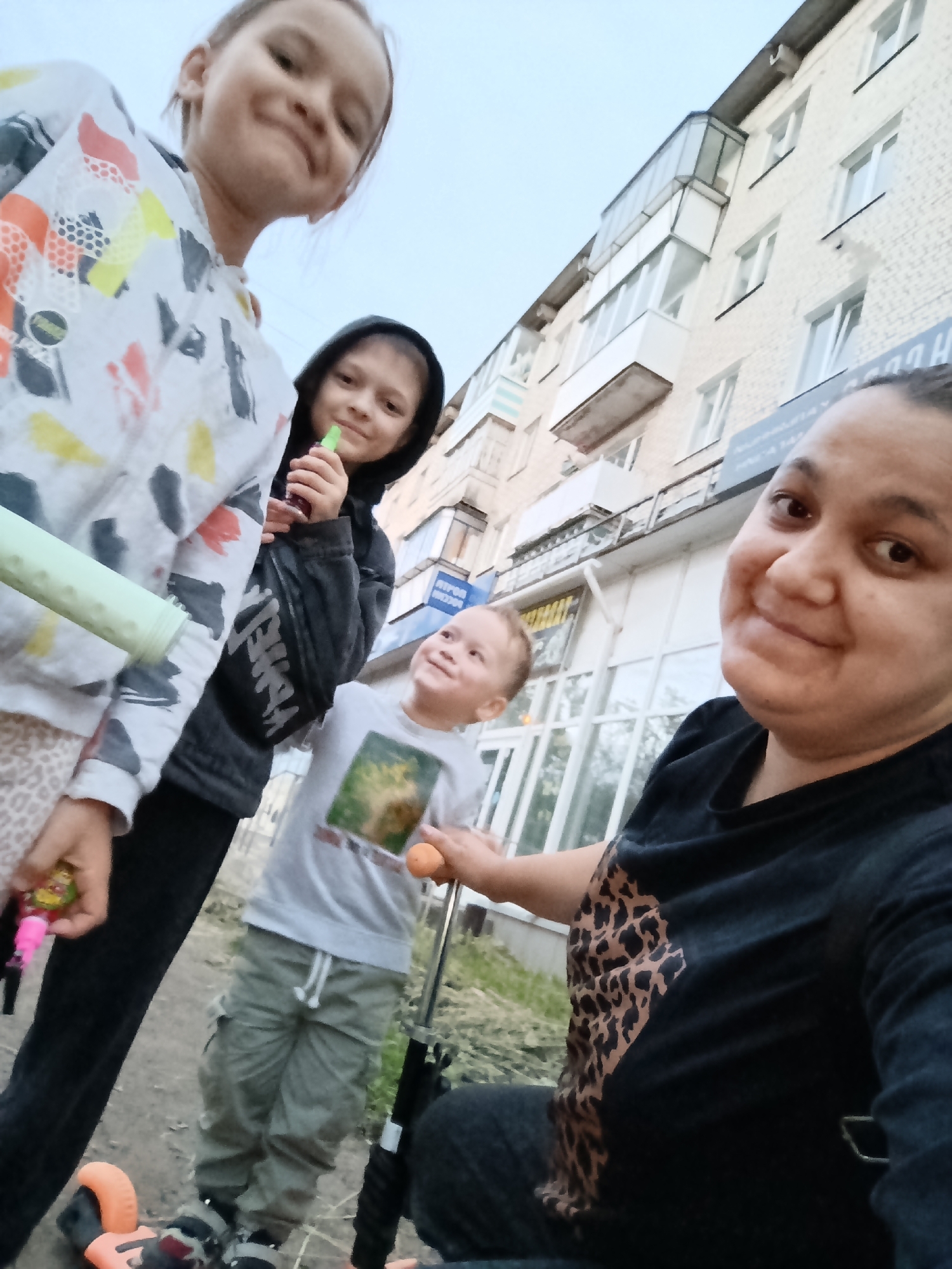 This is a selfie taken outdoors with a group of four people, including three children and one adult. The adult, in the foreground on the right, is smiling and looking at the camera. She is wearing a black t-shirt with a leopard print design on the left side. To her left are three children. The child closest to the adult is a young boy wearing a light grey t-shirt with a picture on it and grey cargo pants. He is looking up and smiling, holding an orange object in his right hand. Next to him is another child, a girl wearing a black hooded jacket, who is holding a green object up to her mouth. To the far left is a third child, a girl wearing a colorful patterned hoodie. She is also smiling and looking towards the camera.
The setting appears to be a residential area, with a multi-story building visible in the background. The building has several windows and a balcony. There is a sidewalk or pathway in front of the building, and some grass and dirt on the ground.
The lighting suggests it is daytime, possibly late afternoon or early evening, as the sky is overcast. The overall mood of the image is happy and casual, with everyone smiling.
There are no visible signs of text in the image.