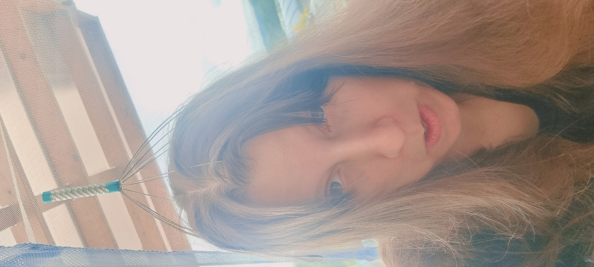 A light-skinned female is seen looking directly at the camera with a head massager on top of her head. The background shows light coming through a window with horizontal blinds covered by a translucent net curtain. The location is Dunnae-myeon, South Korea.