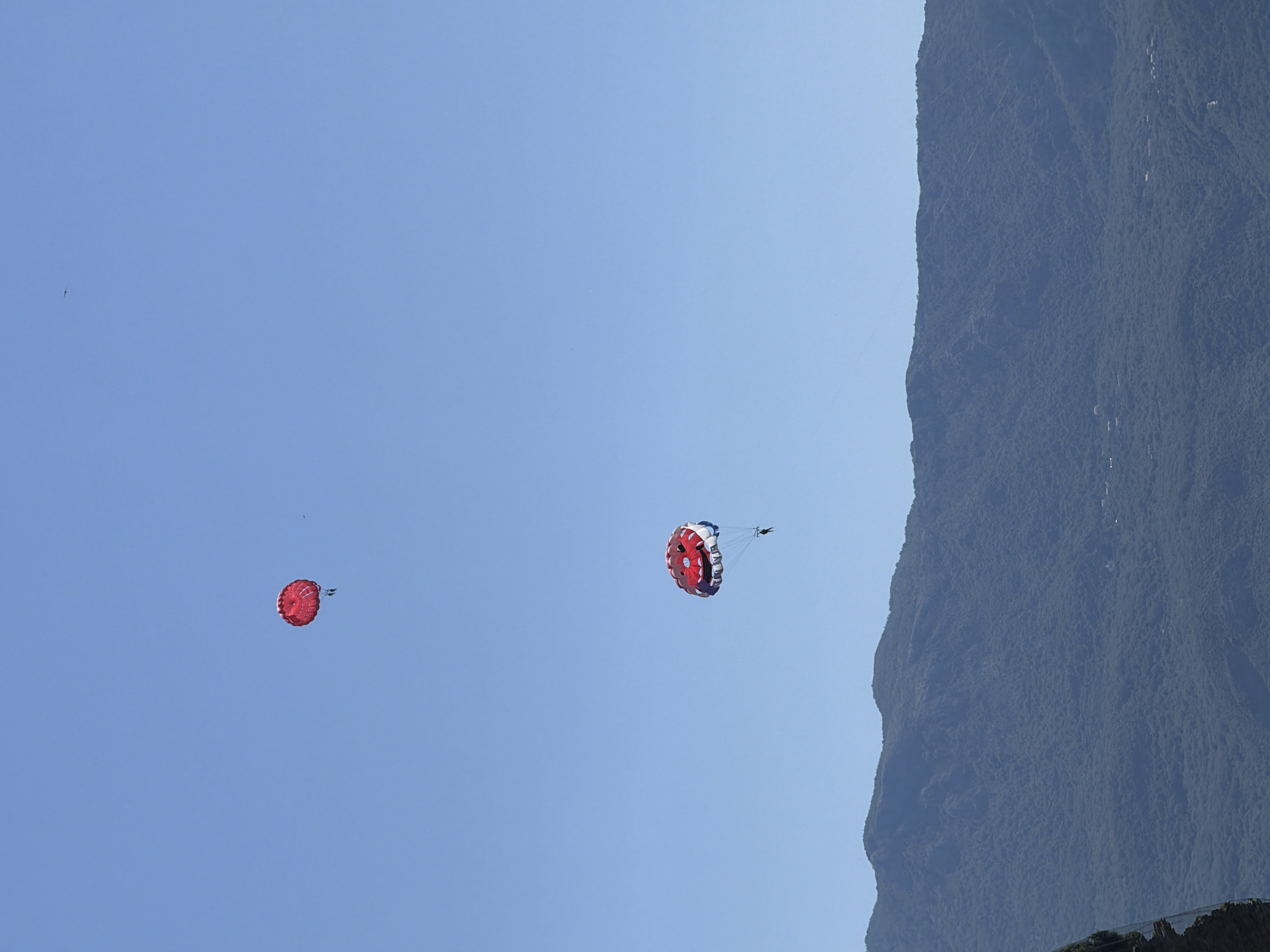 Two parachutes are visible in the sky over the mountainous coastline of Budva, Montenegro. The parachute in the upper portion of the image is solid red and carries one person. The parachute in the lower portion of the image is red and white, shaped like a face, and also carries one person. The sky is clear and blue. The mountains are covered in lush green vegetation and extend to the horizon.