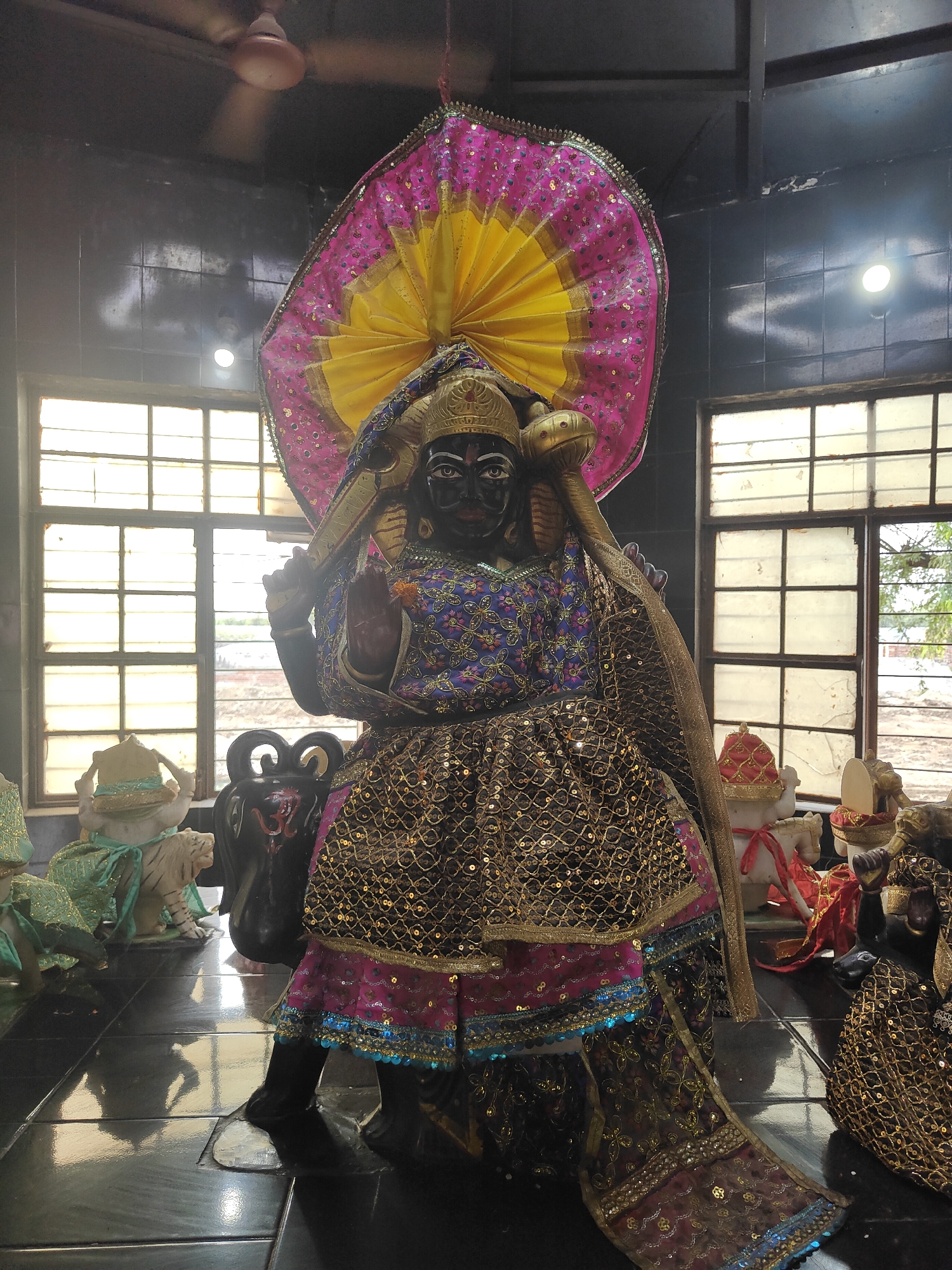 The image depicts a statue of a Hindu deity within a dimly lit room. The statue is adorned with a colorful, ornate costume featuring sequins and metallic threads. It has dark skin and is wearing a headdress that fans out like a halo. Several other smaller statues and objects are visible in the foreground and background, including what appears to be a white tiger statue and a black urn-like object with an "Om" symbol. The room has large windows with a grid pattern, suggesting they might be in a temple or shrine. A ceiling fan is visible in the upper left corner, and the floor is highly reflective, possibly made of polished tile. The lighting suggests artificial light sources within the room. There are no people visible in the image. The style of the statues and decorations is indicative of a religious or cultural site in India. While specific details about Gurgaon are not present in the visual information, such statuary is common in religious establishments throughout India.