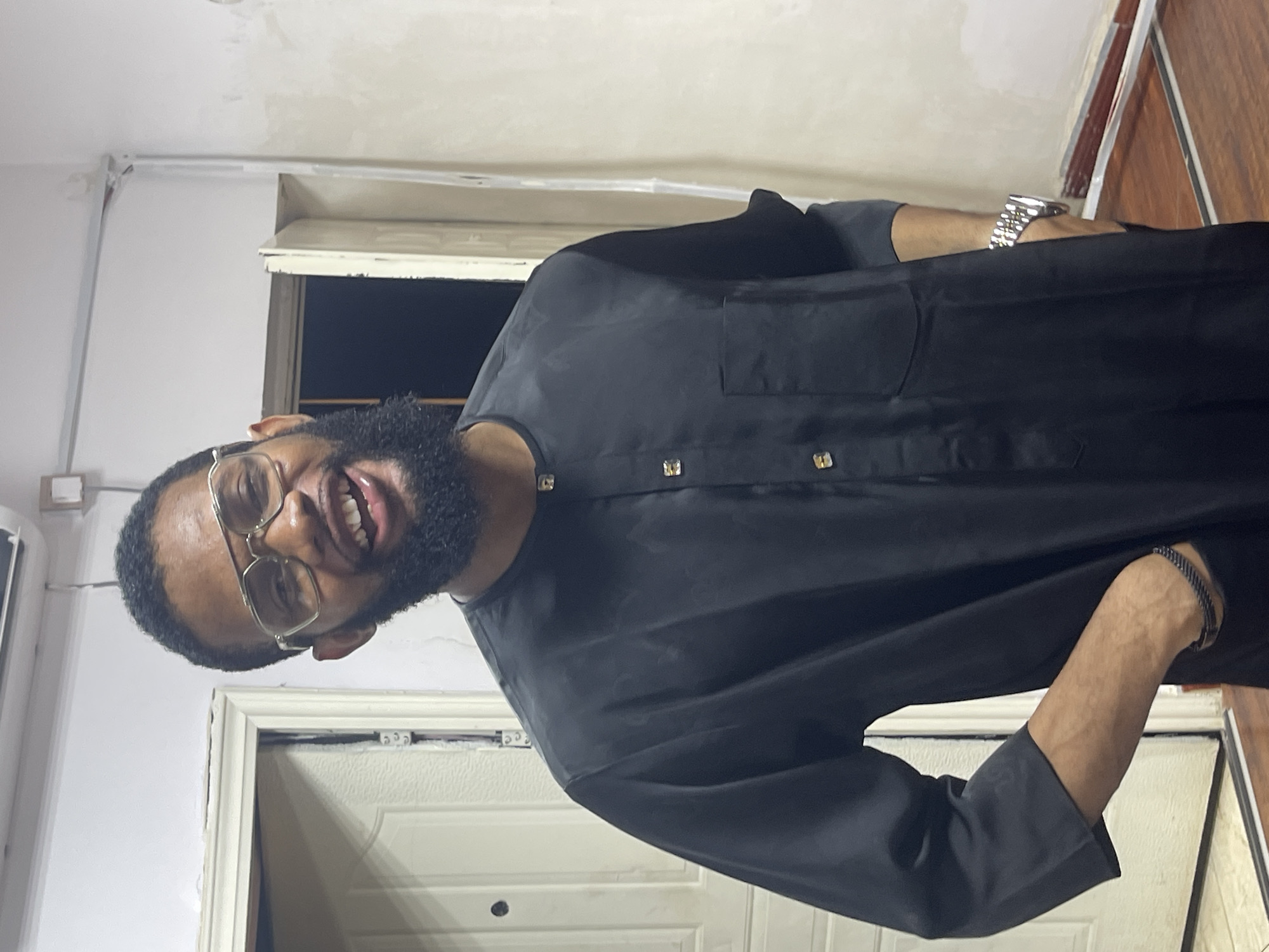 An indoor portrait, originally rotated 90 degrees clockwise, features a man with dark skin, a full dark beard, and gold-rimmed glasses. He is smiling widely, revealing his teeth, and appears to be in a cheerful mood. He wears a dark, long-sleeved traditional garment, resembling a kaftan or agbada top, which is adorned with square gold-colored buttons or embellishments running down the front. On his left wrist, he wears a prominent silver-toned metal watch with a linked band, and a dark, possibly braided, bracelet is visible on his right wrist.
The man's head is tilted, and his upper body is captured from an unusual angle, giving the impression he is looking down into the camera. The background consists of light-colored walls, likely white or off-white, with a white door frame and a closed white panel door. A dark rectangular opening, possibly a window or another doorway, is visible behind his left shoulder. To the right, a section of wooden paneling is present, suggesting a common interior design element in a home or office in Abuja, Nigeria. The lighting is artificial, consistent with an indoor setting, and no visible text is present.