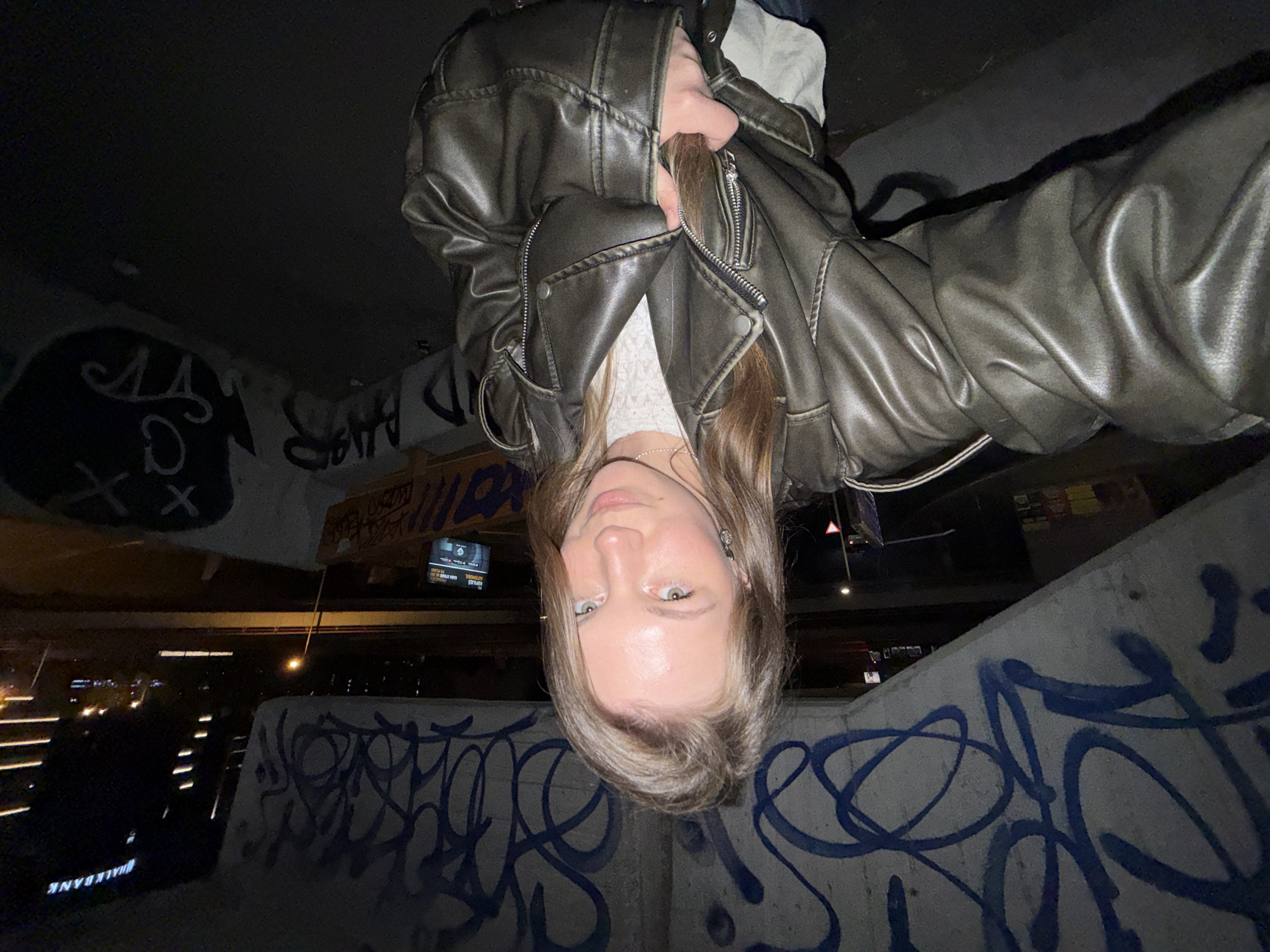 An inverted close-up selfie captures a young woman looking directly at the camera. Her face, with light-colored eyes and fair skin, is centrally positioned and illuminated, suggesting the use of a flash or phone screen. Her light brown hair falls around her head and towards the bottom of the frame (if the image were upright). She wears a dark grey or brown leather-style jacket over a white top, with a delicate necklace visible.
The background, if the image were upright, depicts an urban underpass or bridge at night. Concrete walls and structures are heavily covered in graffiti, predominantly blue spray paint tags, but also some black and white markings, including an 'X' symbol. In the mid-ground and distance, city lights illuminate the scene, showing what appears to be a street or highway with vehicle lights and illuminated buildings.
Notable details include the dark time of day, emphasized by artificial lighting, and the gritty urban environment. Visible text includes "#TALKBANK" on a lighted sign in the lower left (when upright) and partially visible graffiti that appears to spell "BALKANS" in the upper left. A digital display sign in the middle-right background shows white text on a dark screen, including words like "PLANOVI" (plans) and "EVRO", which aligns with the given location context of Belgrade, Serbia.