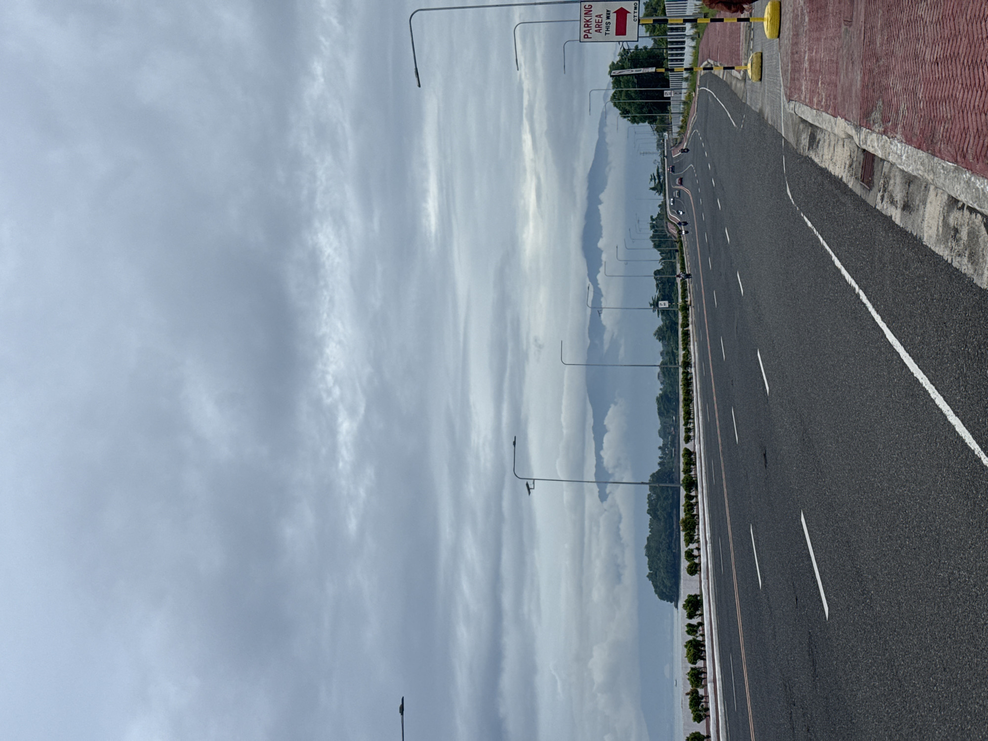 The image shows a road in Davao City, Philippines, under a cloudy sky. The road is asphalt and has white lane markings. There are streetlights lining the road. A sign indicates "Parking Area This Way". Along the right side of the road, there is a sidewalk with red brick-like paving and a concrete curb. There are trees and possibly a body of water visible in the background. Several vehicles are visible in the distance on the road.