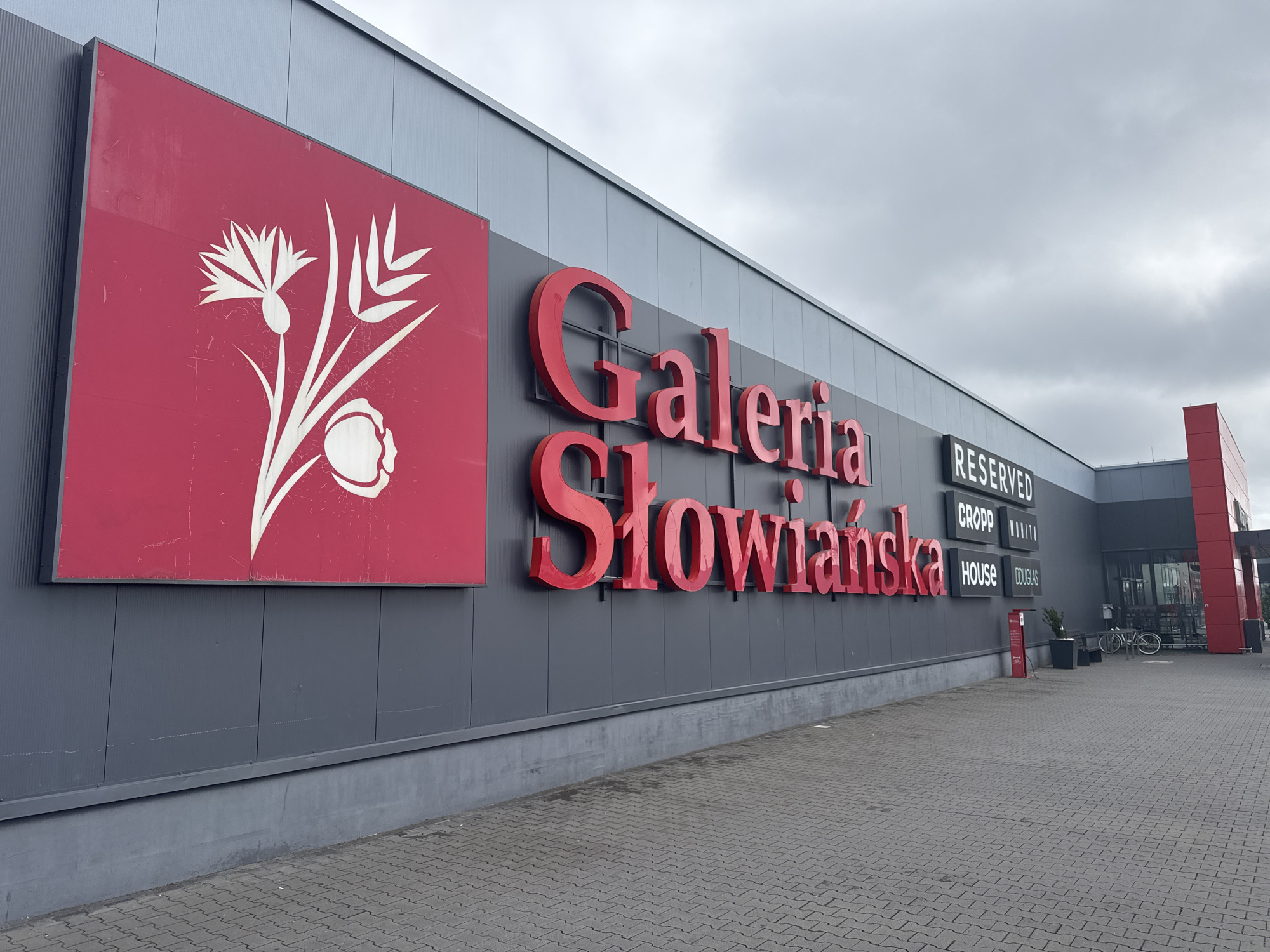The image shows the exterior of the Galeria Słowińska shopping mall in Zgorzelec, Poland. The mall's facade is predominantly dark gray with large red lettering spelling out "Galeria Słowińska". To the left, a large red square sign features a white stylized drawing of a flower and seed heads. Above the main entrance, several smaller signs indicate store names: "RESERVED", "CROPP", "HOUSE", and "DOUGLAS". The ground in front of the mall is paved with gray interlocking bricks. In the background, a bicycle is parked near some planters and a bench, and the entrance to the mall is visible. The sky is overcast.
