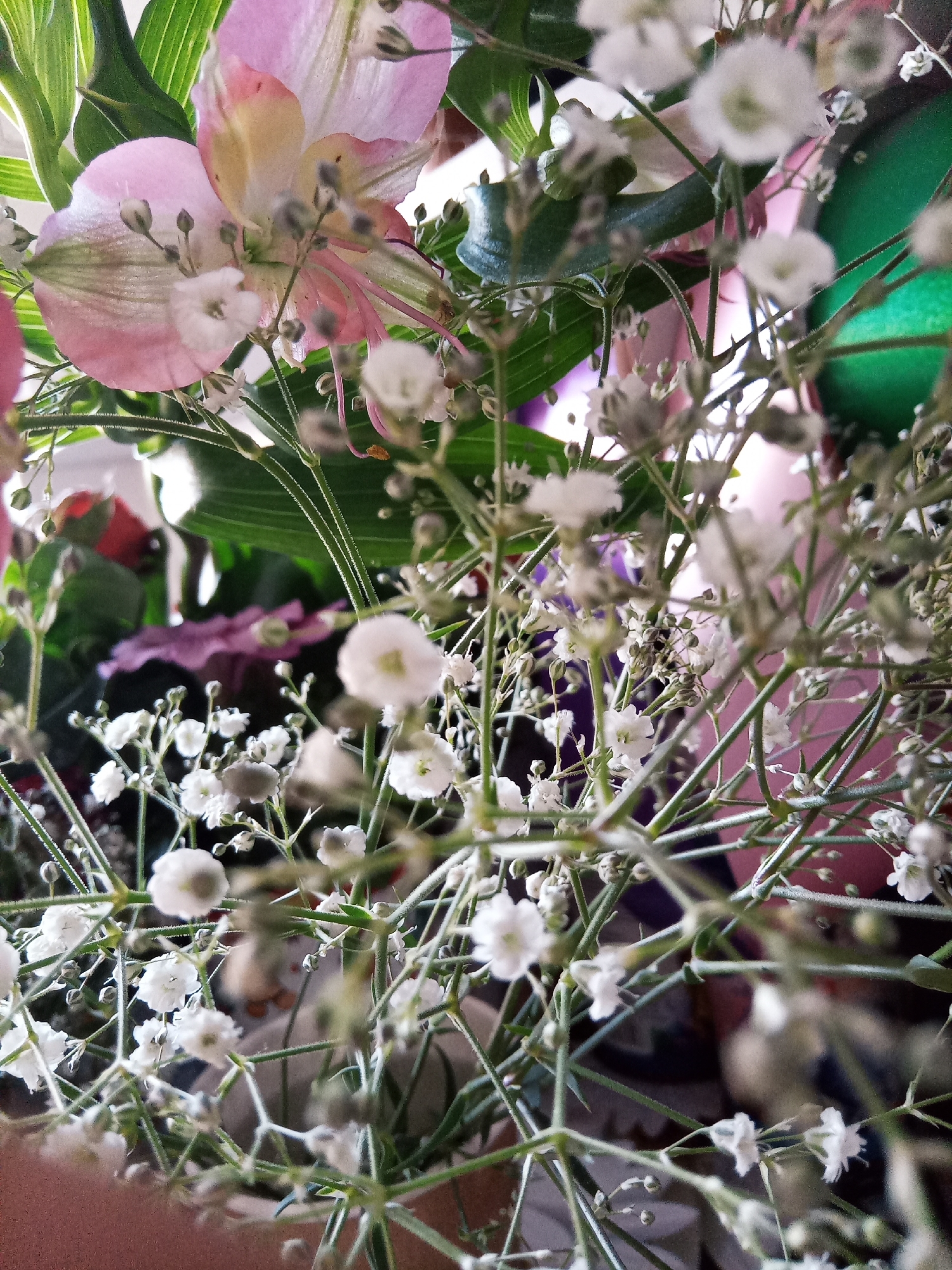 The image is a close-up, shallow depth of field photograph of a floral arrangement. The primary focus is on delicate white baby's breath flowers and their thin green stems, which create a dense, airy network. Behind the baby's breath, larger flowers are visible, including pale pink petals with yellow accents, and some green foliage. The background is softly blurred, with hints of other colors like green, pink, and purple, suggesting other elements within the arrangement or the surrounding environment. No specific location in Chișinău, Moldova, can be identified from the visual information provided. There are no people or actions depicted in the image.