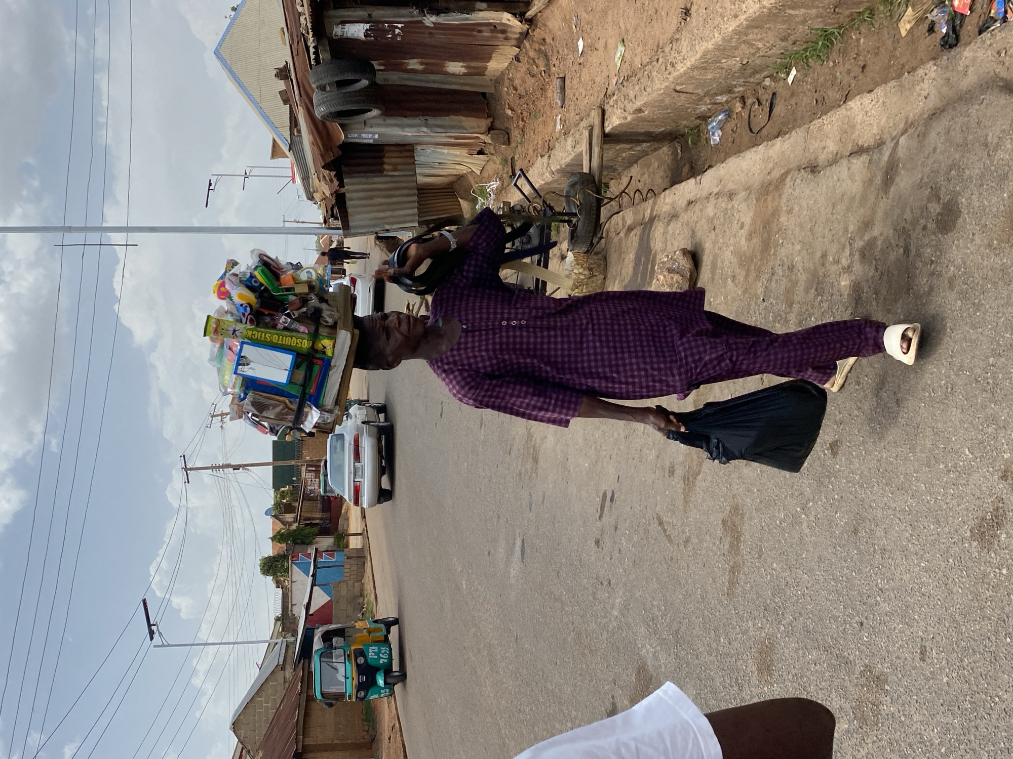 A man walks on a street in Jos, Nigeria. He carries a collection of household goods balanced on his head, and a black bag in his right hand. He is wearing a purple, patterned, long-sleeved shirt and pants with white slippers. A vehicle is parked to his left, and a green and yellow auto-rickshaw is further down the road. Buildings made of corrugated metal sheeting and concrete line the street, and power lines cross the sky.