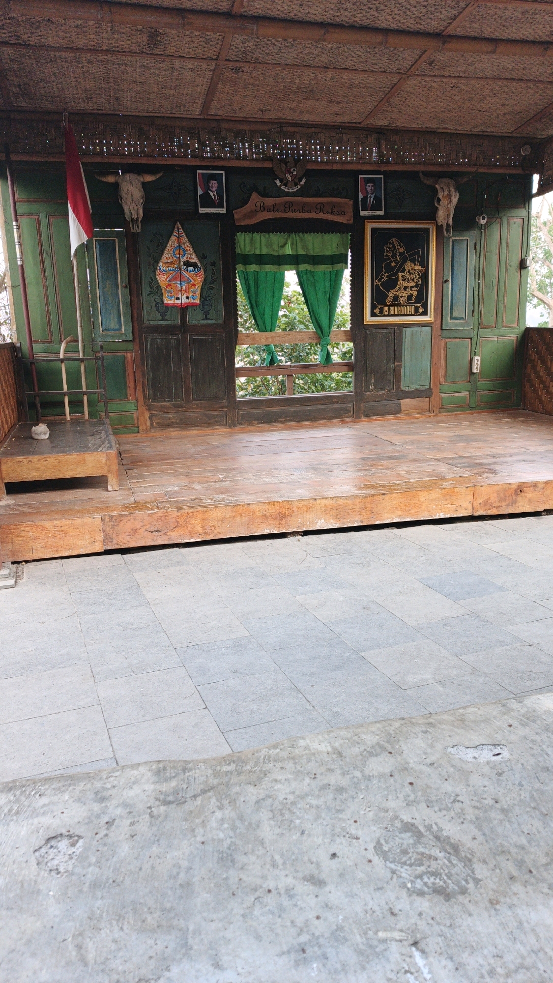 The image depicts an outdoor stage or performance area with traditional Indonesian elements, located in Pojok, Indonesia. The stage is constructed of weathered wooden planks, elevated from a paved area in the foreground. Behind the stage, a backdrop features dark green wooden panels, adorned with various cultural artifacts. These include a bull's skull on the left, a Wayang puppet illustration, portraits of men in formal attire, and a framed depiction of a traditional Indonesian figure. A wooden sign above the green curtains reads "Bale Purba Reksa." A red and white Indonesian flag hangs from a pole on the left side of the stage. The structure is roofed with woven natural materials. Trees and foliage are visible through the opening behind the curtains, suggesting an outdoor setting. The overall impression is one of a cultural performance space, possibly a community gathering point or a venue for traditional arts.
