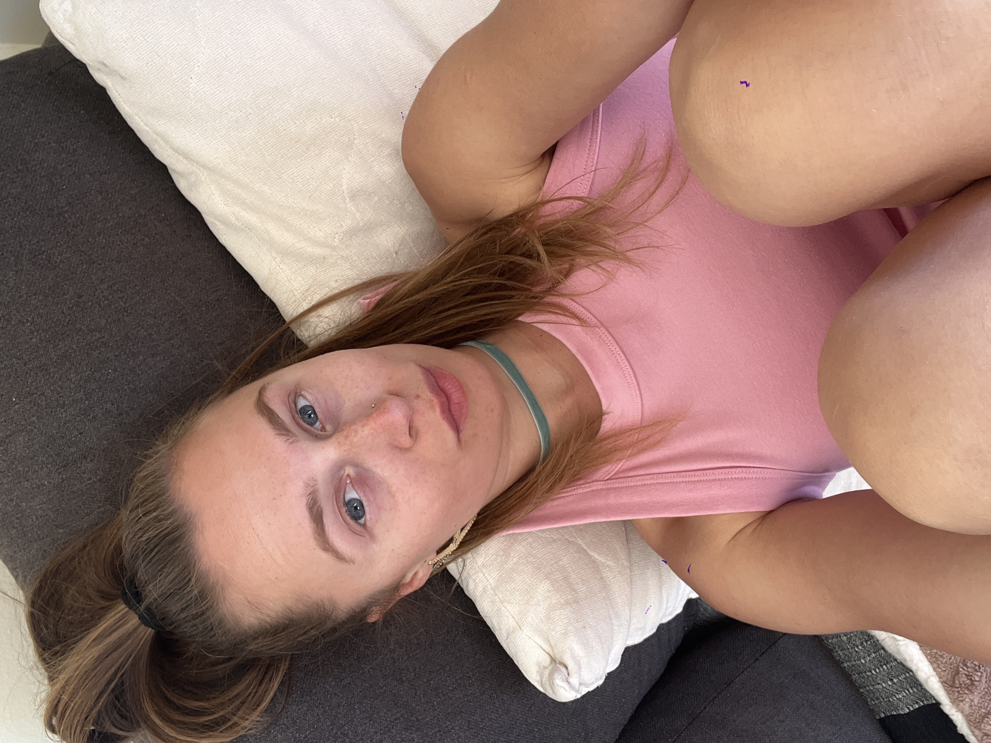 A young woman is laying on a grey couch with a white pillow. She is wearing a pink tank top and a green choker. She has fair skin, freckles, and blue eyes. Her hair is brown and pulled back in a ponytail. She is wearing earrings. The location in the city of Bečići, Montenegro, cannot be determined from the image.