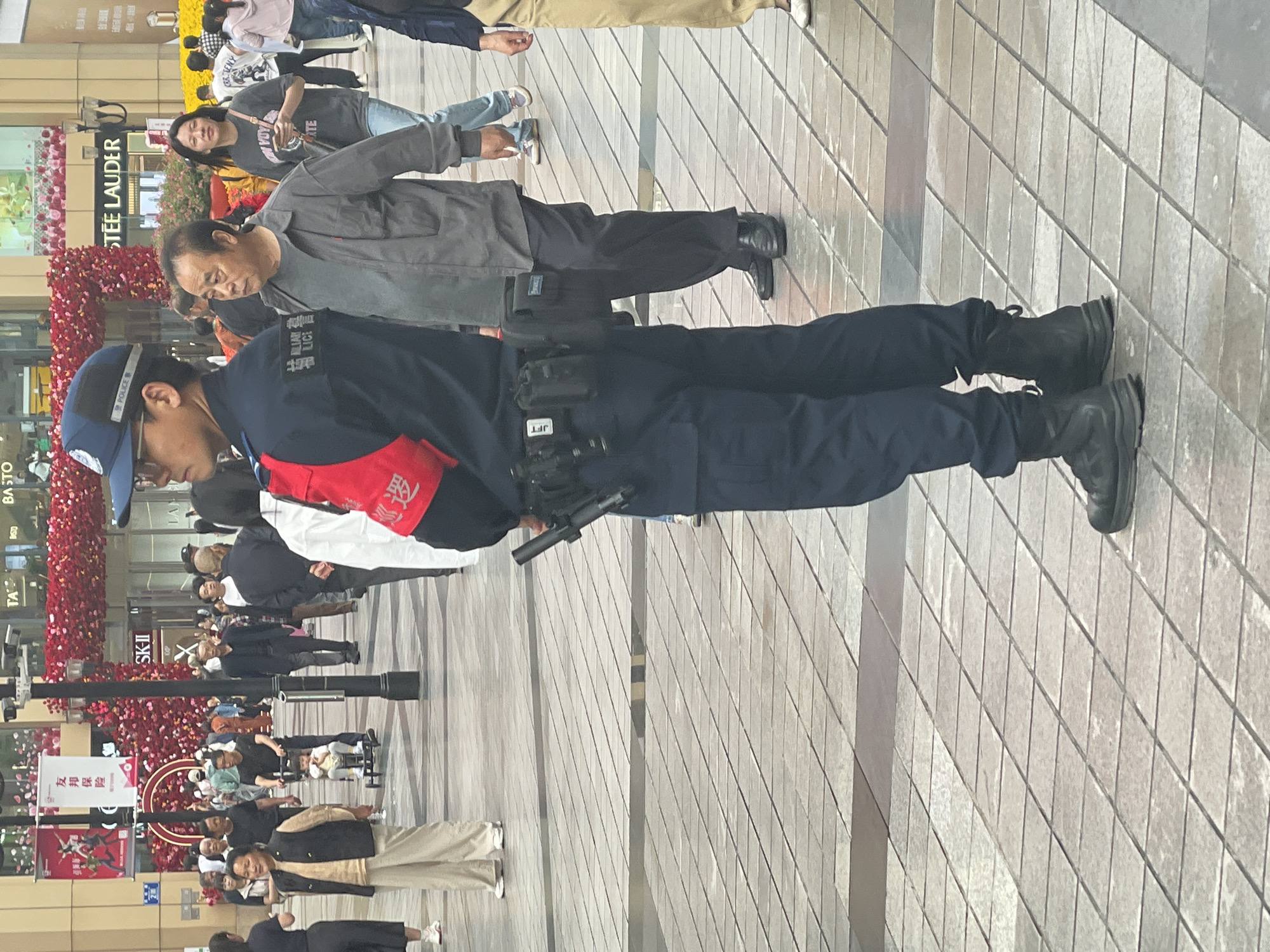 The image shows an outdoor scene, likely a pedestrian area in Yuzhong District, China. A police officer in a dark blue uniform, wearing a cap with "POLICE" on it, stands with a pistol holster on his belt. A red armband with Chinese characters is on his left arm. He is standing on a tiled surface near a man in a gray shirt. In the background, there are storefronts, including "STEE LAUDER," and decorations of red flowers. People are walking in the background. Signage indicating "友邦保险" (AIA Insurance) and "SK-II" can be seen.