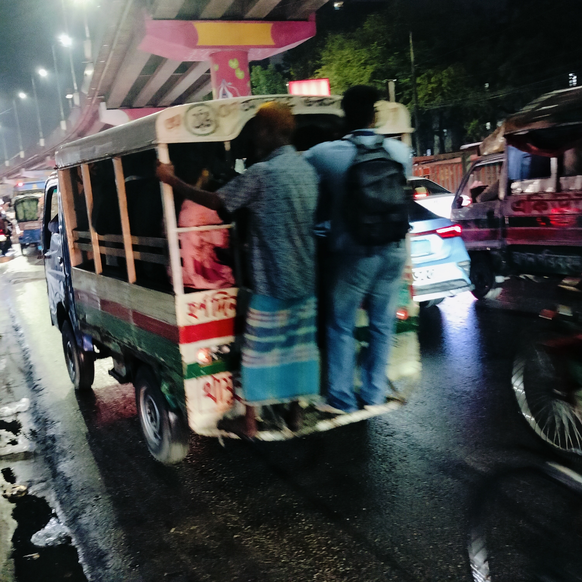 The image depicts a nighttime scene in Dhaka, Bangladesh, characterized by wet roads reflecting ambient light, indicative of recent rain. The focus is on a "tempo," a type of three-wheeled public transport vehicle, moving through traffic. Two men are standing precariously on the tempo's rear platform, holding on for support. The man on the left is wearing a patterned shirt and a lungi, a traditional garment. The man on the right has on a blue shirt, jeans, and a backpack. Other vehicles, including cars and another tempo, are visible in the background, along with trees and an elevated structure, possibly an overpass, with pink and yellow accents, a common feature in Dhaka's urban landscape.