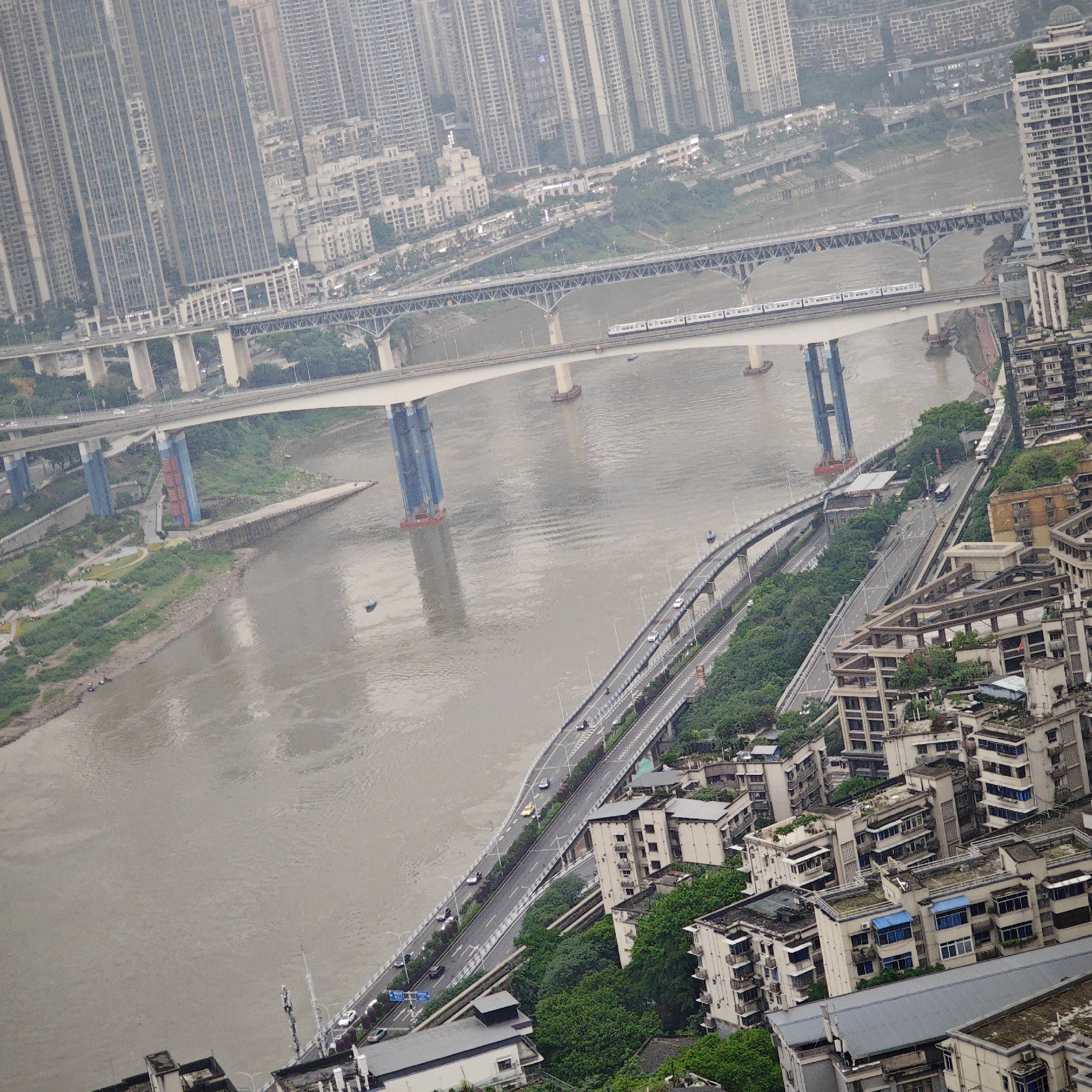 The image shows an aerial view of Yuzhong District, China, featuring a broad, murky river spanned by several bridges. The bridges appear to carry both vehicular and railway traffic, with a train visible on one. High-rise buildings dominate the cityscape in the background, shrouded in a haze. The riverbanks are partially covered in greenery, and roads run alongside the river, lined with vehicles. The overall impression is of a densely populated urban area with significant infrastructure for transportation. Buildings with blue-roof tiles are seen in the lower right foreground.
