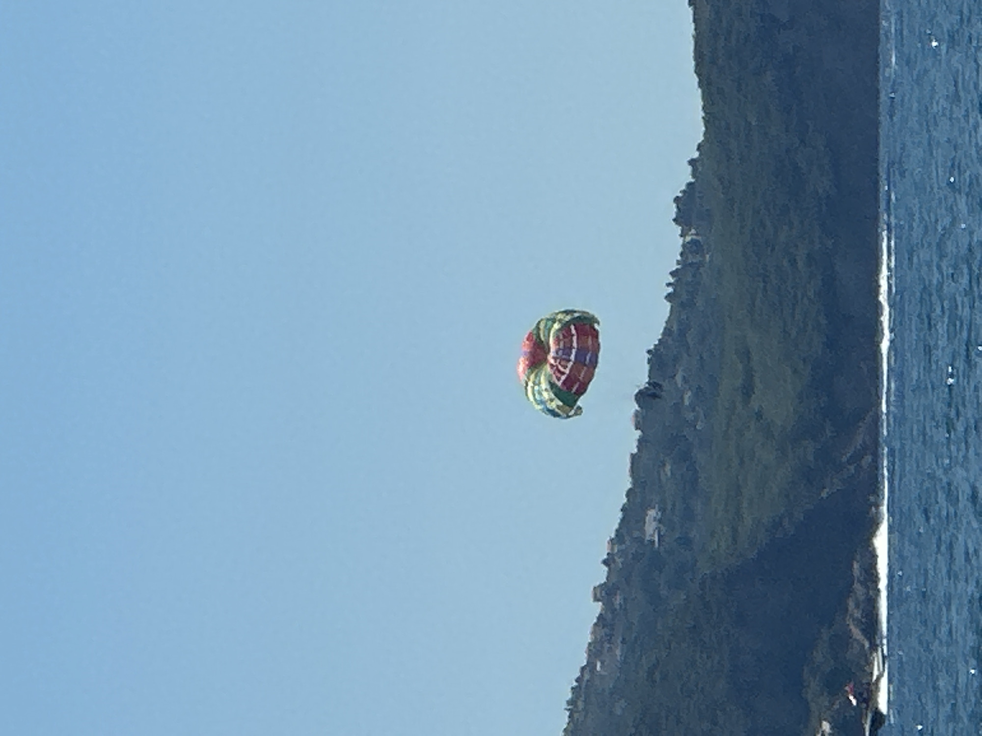 A colorful parasail is suspended in the air against a clear blue sky over a body of water. Below the parasail, a lush green hillside is visible, dotted with houses. The water in the foreground is a deep blue with small whitecaps, indicating gentle waves. The scene appears to be a coastal area, possibly in Boreti, Montenegro, during daylight hours, suggesting a recreational activity such as parasailing is taking place.