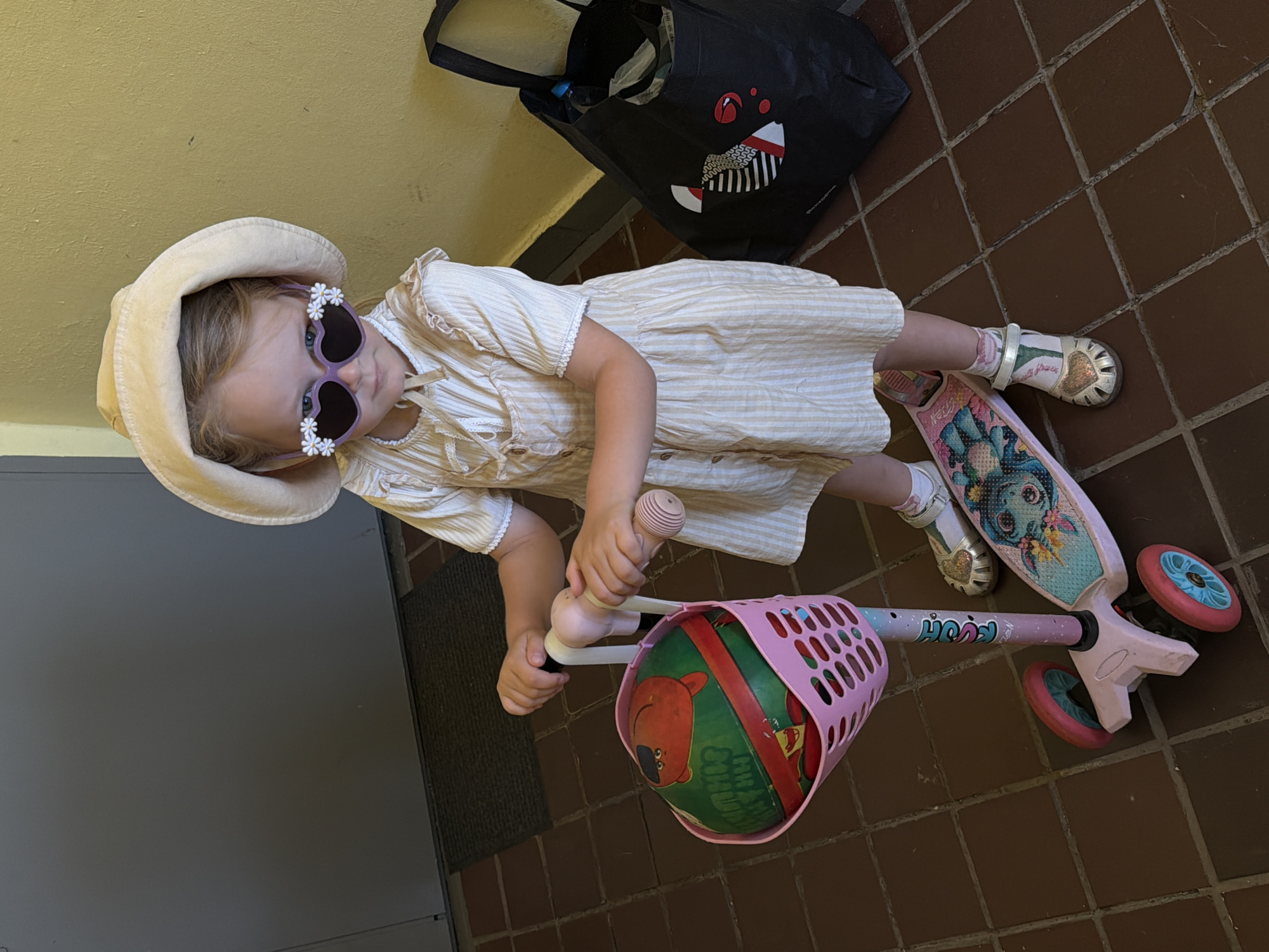In a residential building hallway in Khimki, Russia, a young girl stands on a pink three-wheeled scooter. She wears a light-colored, striped dress, a matching sun hat, and heart-shaped sunglasses adorned with white flowers. Her feet are clad in white socks with a floral pattern and gold Mary Jane shoes. A pink basket on the scooter's handlebars holds a green ball with red and black cartoonish figures. To the right of the girl, a black tote bag with a stylized face design rests against a yellow-painted wall. The floor is tiled with dark brown square tiles.
