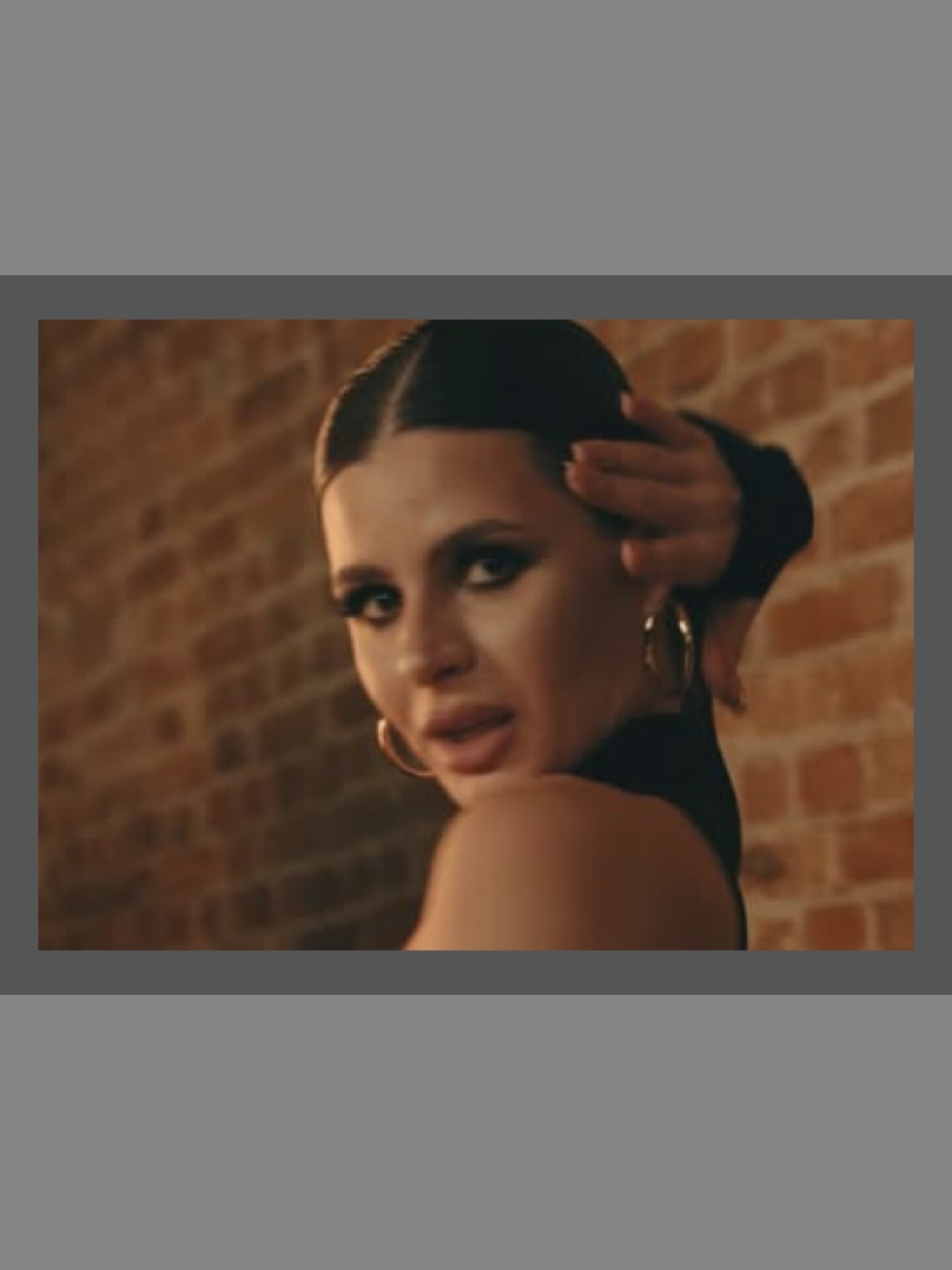 In the foreground of the image, a woman is captured in a close-up shot, her face turned to the side and slightly upward. She has dark hair pulled back, with strands framing her face. Her eyes are made up with dark eyeliner and mascara, and she is wearing large hoop earrings. Her mouth is slightly open, and one hand is raised to her head, with fingers touching her hair. She is wearing a dark, off-the-shoulder garment. The background is a brick wall, illuminated by warm, low lighting. The overall scene appears to be indoors, possibly a studio setting or a dimly lit room. The exact location within a city is not discernible from the image.
