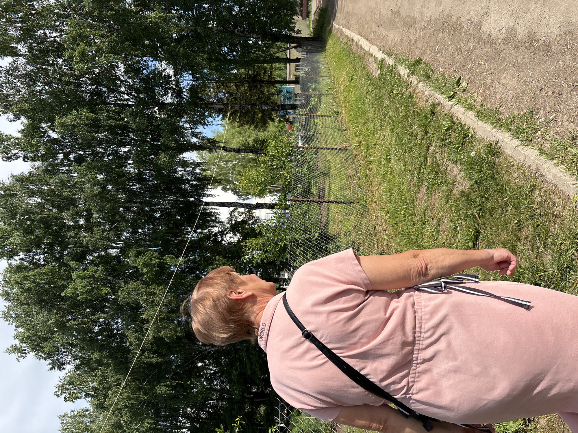 An adult female, viewed from the rear, walks away from the camera on a dirt path in a rural setting. She has short, light brown hair and is wearing a light pink collared shirt with a black strap from a bag across her body. Her shirt has the word "WORLD" embroidered on the collar. She is walking next to a grassy area bordered by a chain-link fence. Behind the fence, there are trees, some buildings, and a blue sign. The path curves to the right and continues into the distance. The sky is cloudy.