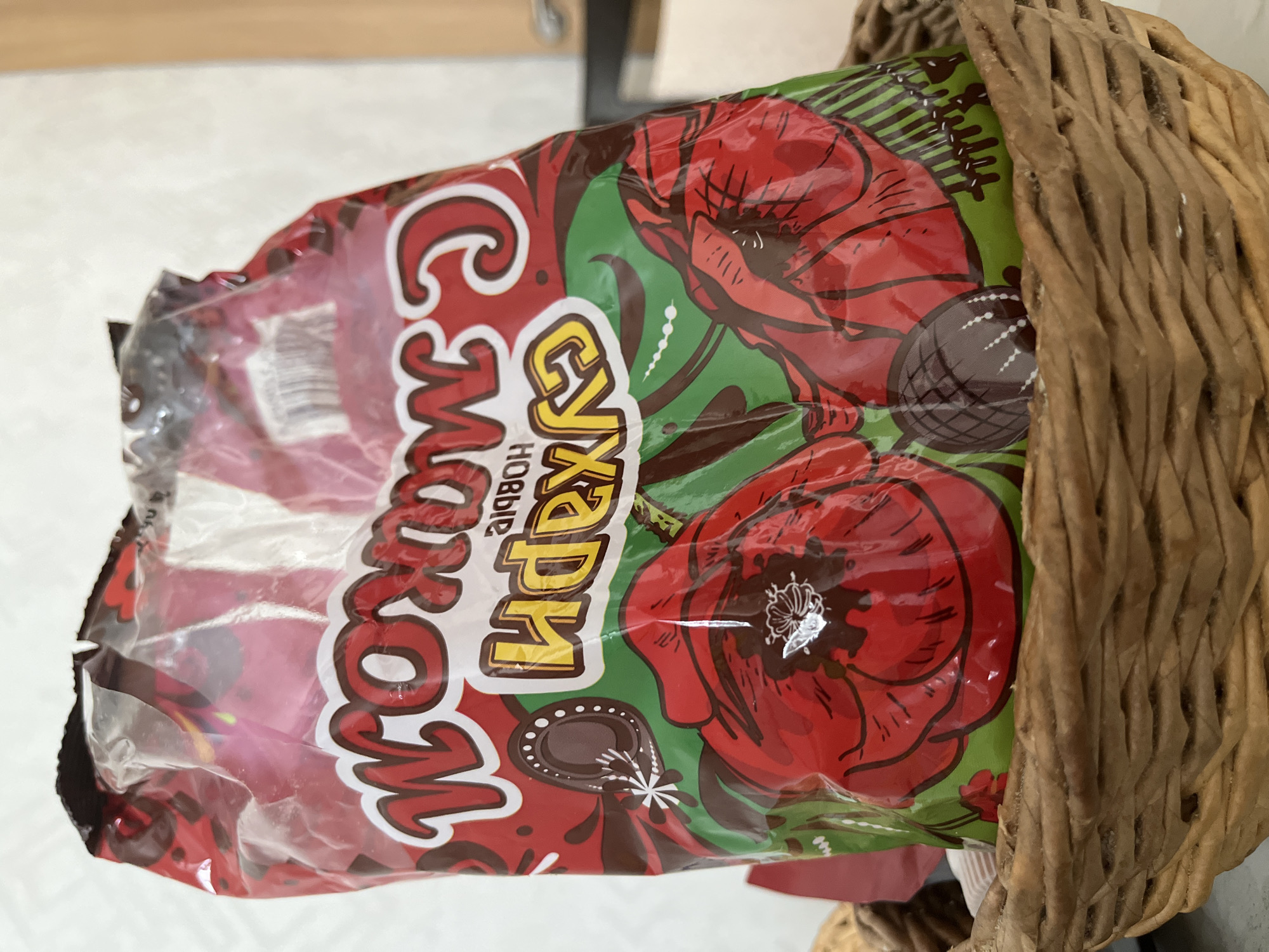 A plastic bag of chocolates is displayed in a woven basket. The bag is red with a repeating pattern of red poppies and green leaves. The text on the bag is in Cyrillic script and appears to be "Шоколадные идеи" which translates to "Chocolate Ideas". A single chocolate truffle is visible peeking out from the bag and another is partially visible in the basket below. The basket is made of woven straw and sits on a flat surface that is out of focus. The background is a plain, light-colored wall with a sliver of a wooden door frame visible on the right side of the image.
