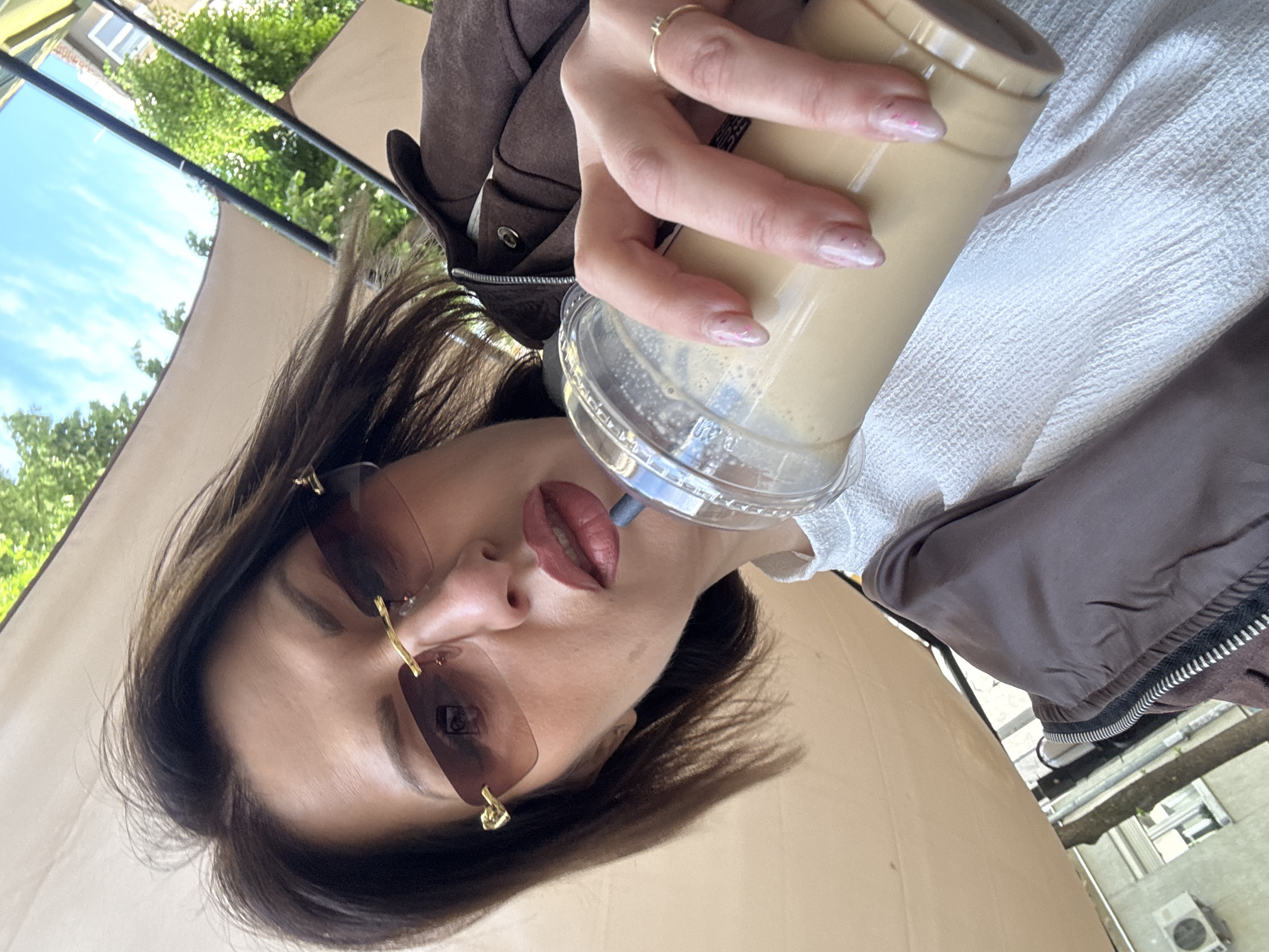 A woman is holding a beverage in a clear plastic cup with a lid and straw. She is wearing sunglasses with gold accents, a brown jacket, and a white top. She also has a ring on her finger. The photo appears to be taken outdoors under an awning, with trees and a building visible in the background. Based on architectural clues and environmental context, the photo might have been taken in Sofia, Bulgaria.