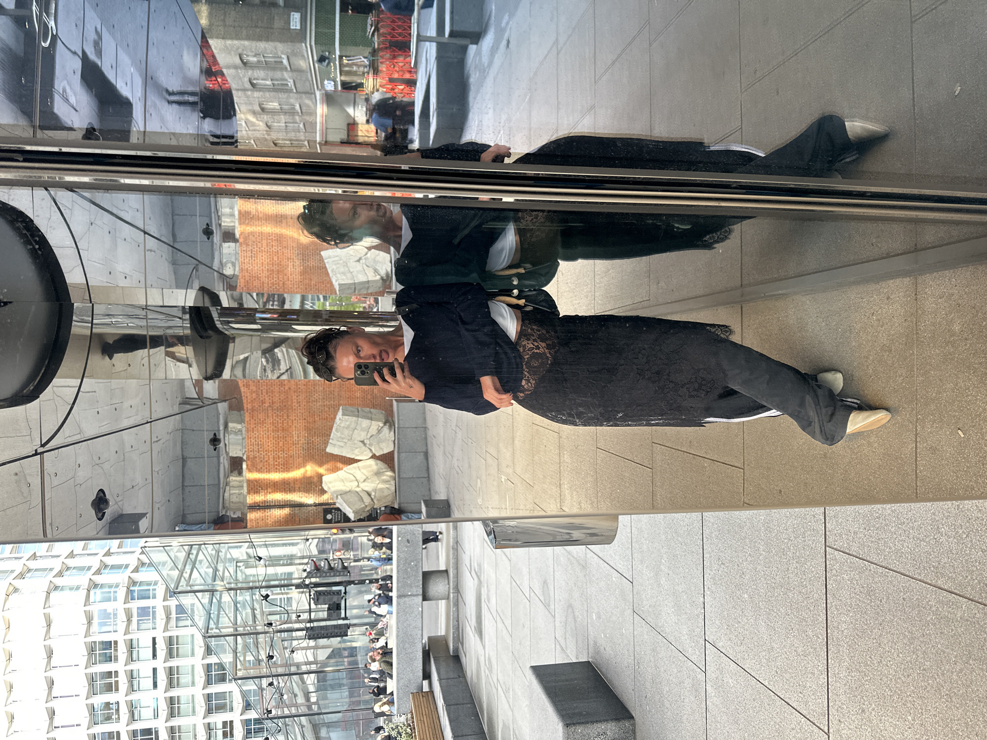 A woman takes a selfie in a reflective surface in a public space. The woman is wearing a black lace dress with a black blazer, a white top, and dark pants, and appears to be standing on a paved surface. Her reflection can be seen above her and the sky is partially reflected as well. The reflection above also shows a brick building. In the distance, there is a partially visible building with a glass roof, an array of scaffolding, and people. The scene is outdoors during the day. Based on the style of the buildings, this is likely a location in the city of London, United Kingdom.