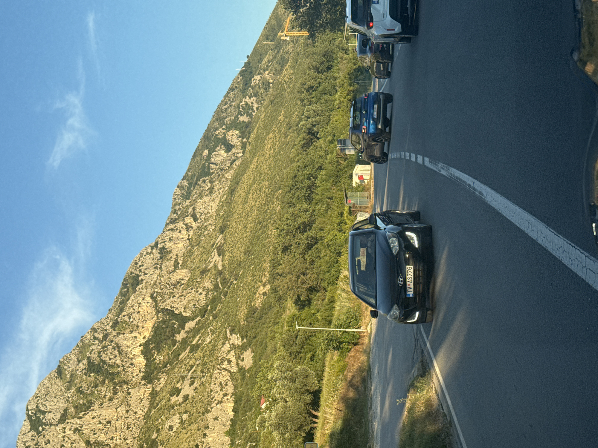 The image captures a view from a vehicle traveling on a road in Blizikuće, Montenegro. A black Hyundai i20 is in the foreground, with its license plate reading "TV AS776". Further down the road, a blue SUV is visible, followed by other vehicles. The scene is set against a backdrop of a steep, rocky mountain covered in green vegetation, with a clear blue sky above. A construction crane is also visible in the distance. The lighting suggests it is either early morning or late afternoon. There are no people visible outside the vehicles.