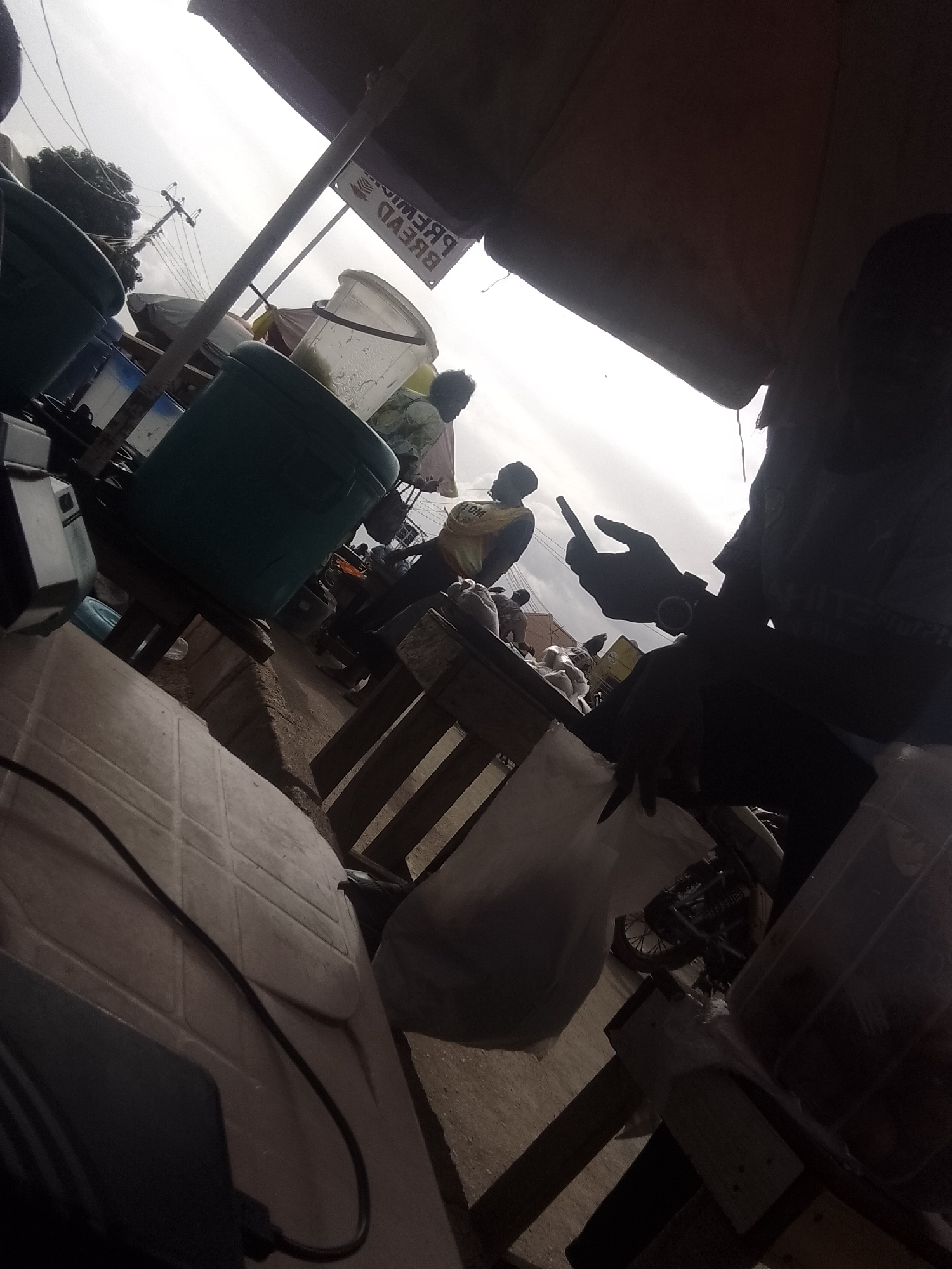 The image depicts an outdoor market scene under a large awning. In the foreground, a person is silhouetted against the bright sky, holding a smartphone. To the left, a large teal container and other containers are visible. Further back, another person wearing a yellow vest is seen. The ground appears to be dirt and some paved surfaces. Overhead, a power line and utility pole are present. The overall atmosphere is that of a bustling marketplace.