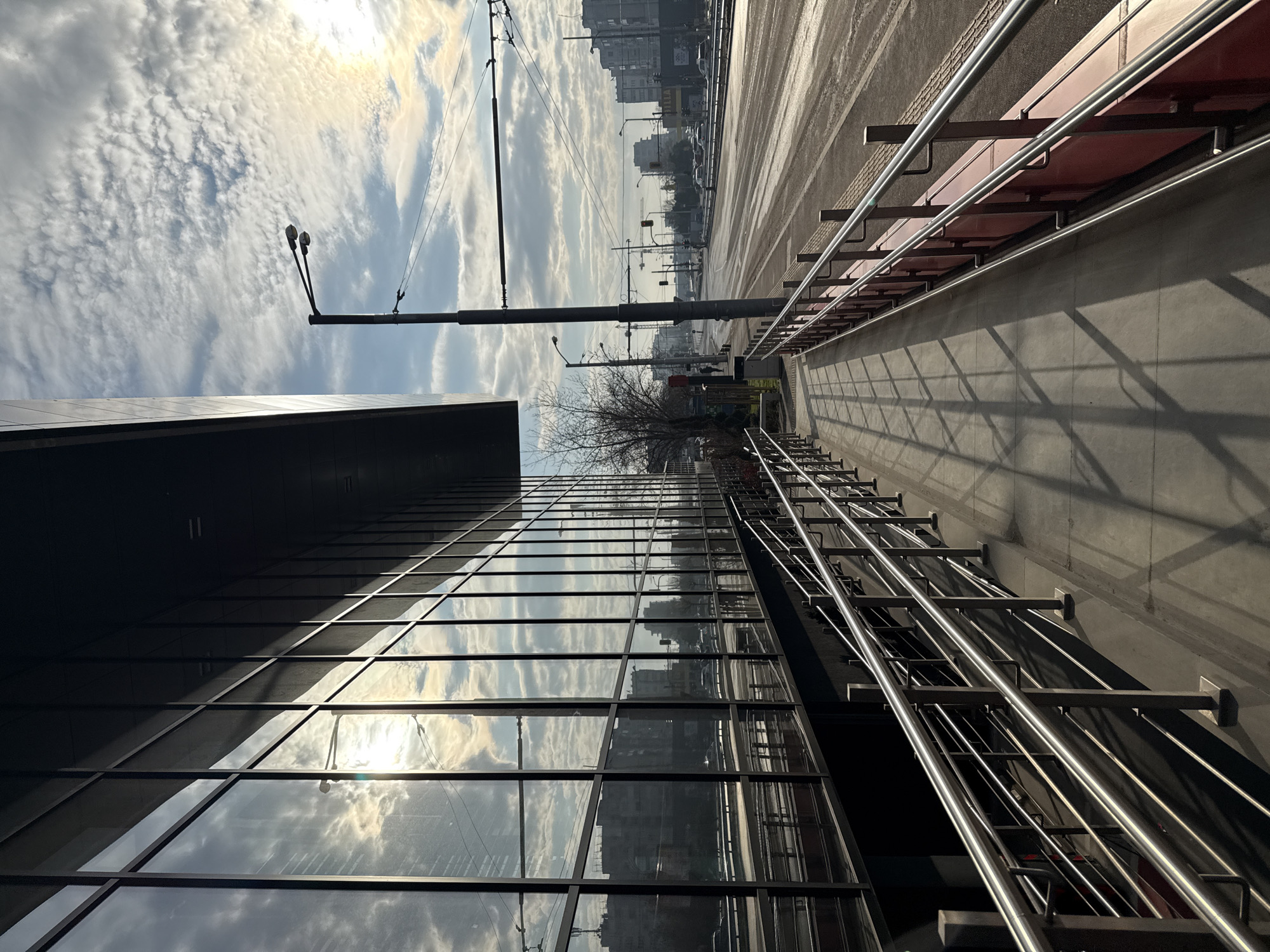 This is a street scene in Sofia, Bulgaria, likely taken near a modern building with a mirrored glass facade. The view is angled upwards, capturing the building's reflection of the sky, which is partly cloudy with a visible sun. A sidewalk or platform is present to the right, bordered by metal railings. Overhead electrical wires are visible against the sky, and streetlights are spaced along the road. In the background, other buildings and possibly construction cranes are discernible. No people are prominently featured in the frame. The overall impression is one of an urban setting with modern architecture.