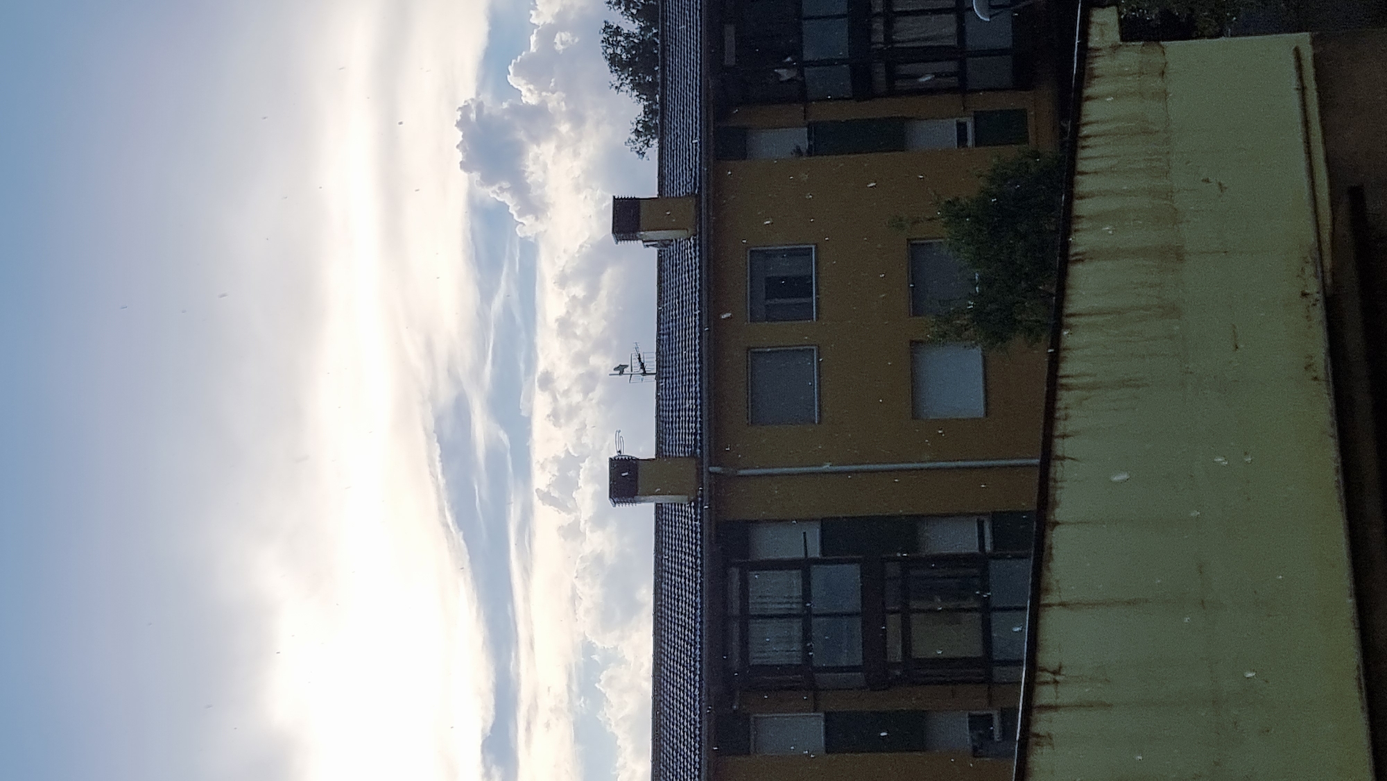 The image depicts a yellow apartment building in Sant Joan Les Fonts, Spain, under a cloudy sky. Small white specks are visible, appearing to be rain or snow. The building has a dark tiled roof and several windows, some of which are dark or reflective. There are two chimneys on the roof, one with an antenna. A greenish-yellow wall with rust streaks runs along the bottom of the frame, partially obscuring the building's lower floors and windows. Trees are visible to the right of the building. The overall scene suggests a potentially inclement weather day.