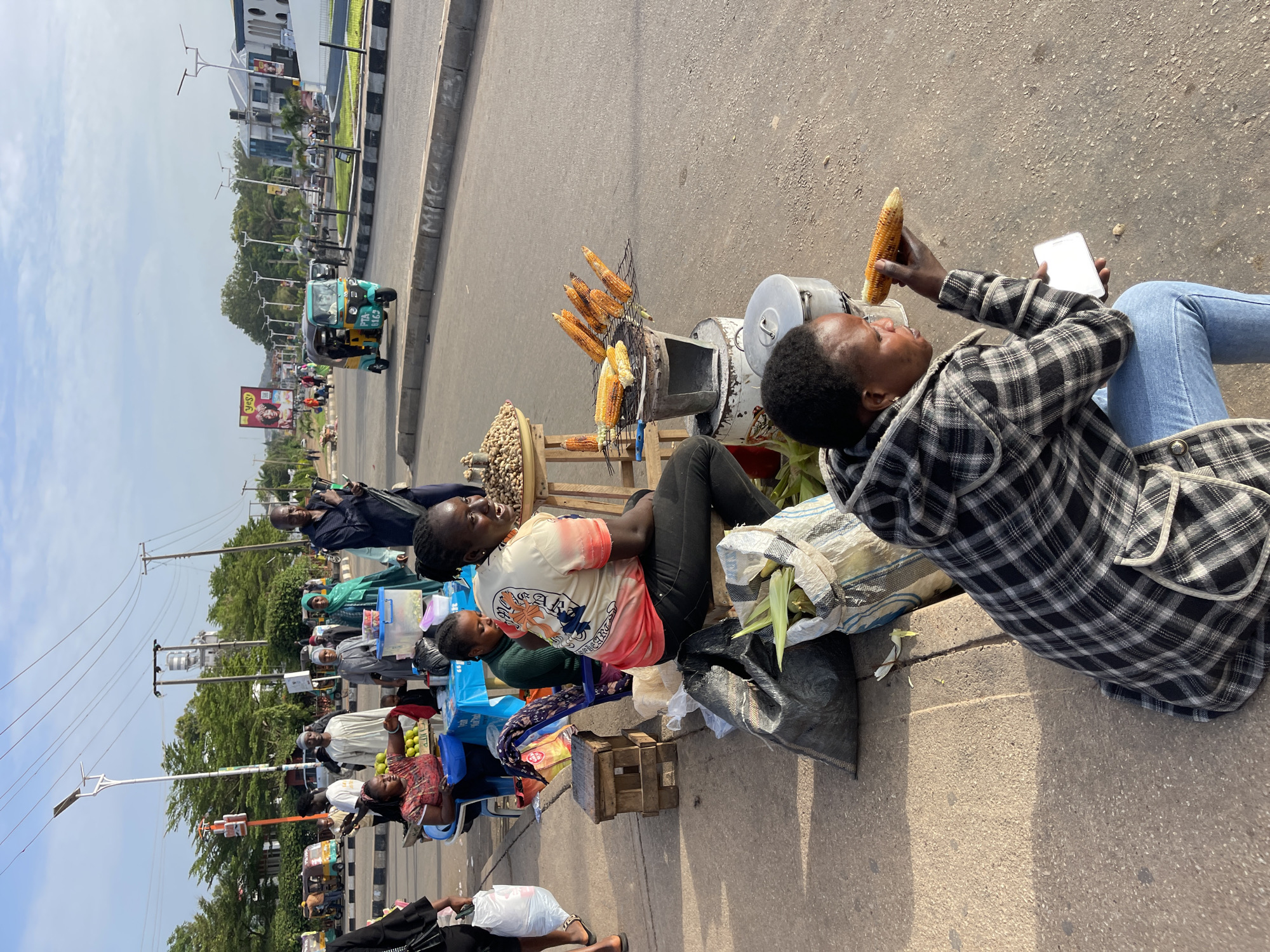 A wide, outdoor, daytime shot shows a street scene in Jos, Nigeria. The horizon is level, showing a clear sky. A road runs from right to left in the foreground, with street vendors along the side. Vendors are sitting on low stools or directly on the ground. A woman in a plaid shirt and jeans is seated, eating corn and holding a smartphone. Another vendor in a t-shirt sits nearby, looking at her. Behind them is a charcoal grill with roasted corn, a pot, and a basket of peanuts. In the background, more vendors are selling various items. Beyond the vendors, there is a road with traffic, including a tricycle taxi. Buildings and trees are visible in the distance. The overall impression is of a busy street scene with informal commerce.