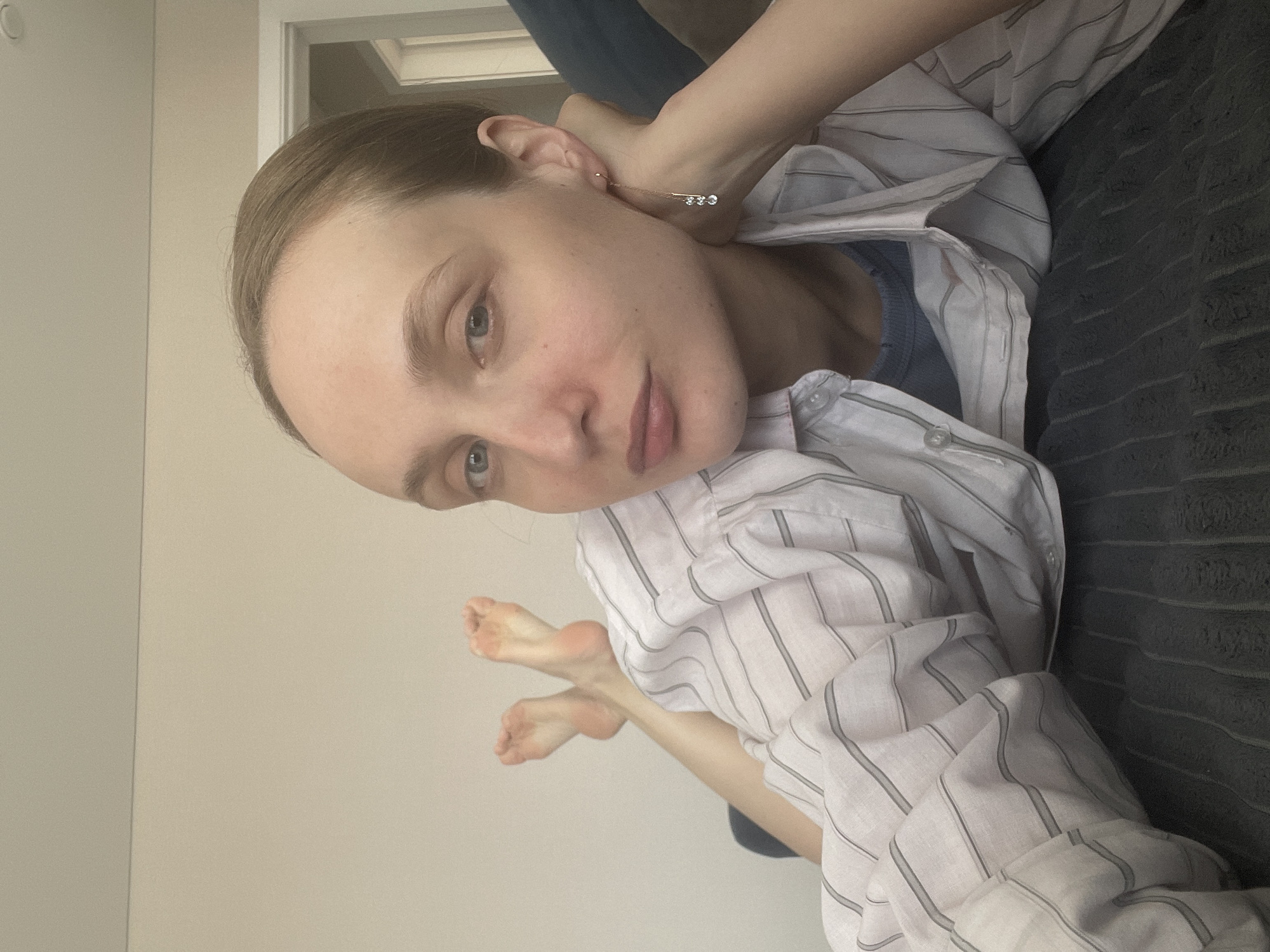 A person lies on a dark gray textured surface, possibly a sofa or bed. They wear a white, vertically striped button-down shirt over a blue undershirt. Their skin appears pale, and they have short, light brown hair. They are wearing a delicate, silver-toned earring with three small, clear stones. Their feet are visible, angled towards the camera. The background consists of a light beige wall and a white door frame. The image is taken at an angle, with the person's head positioned towards the top right corner and their feet toward the bottom left. The location mentioned, Sepych, Russia, does not appear to affect the scene's presentation.