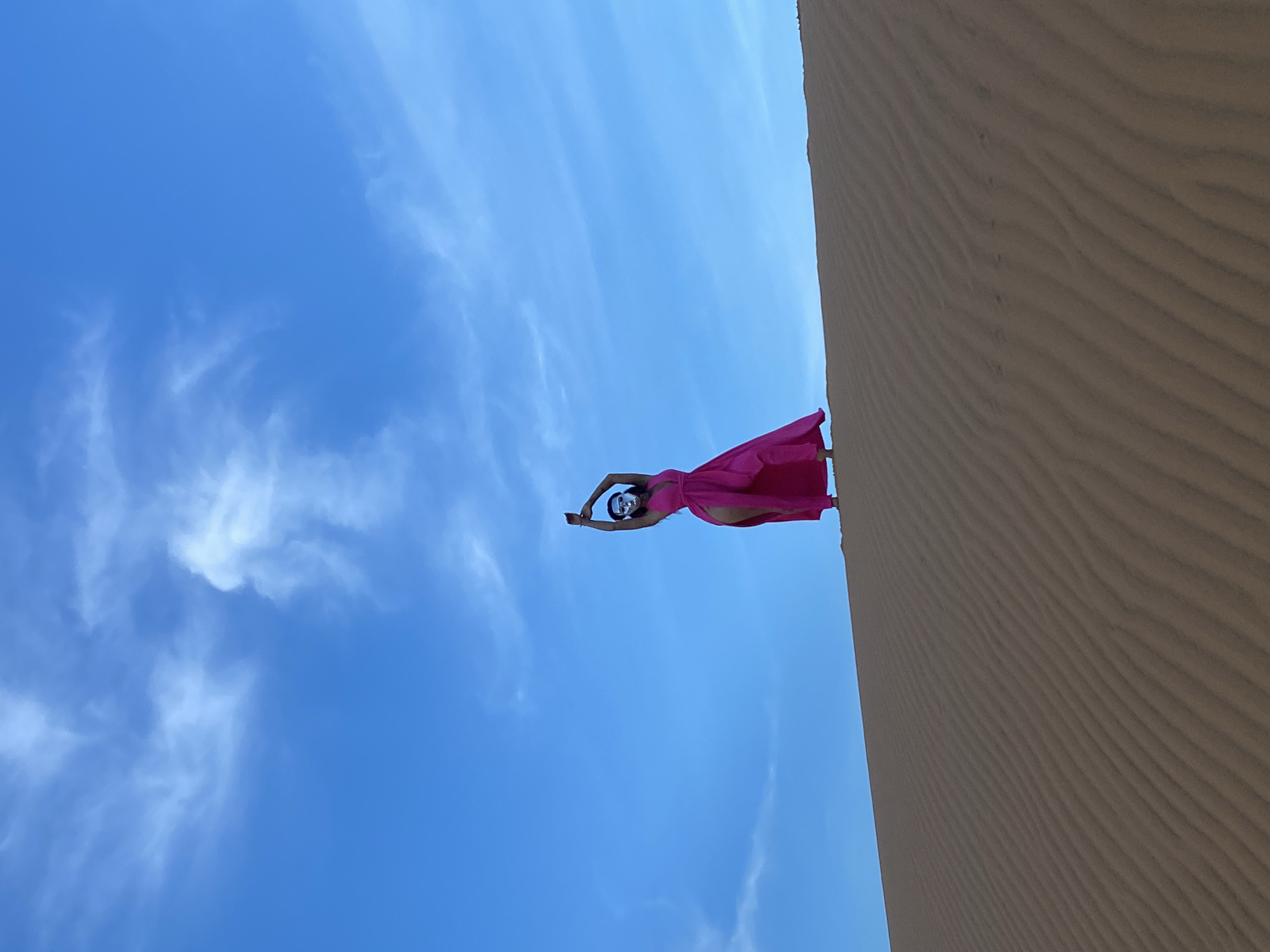 A person in a vibrant pink dress stands on a sand dune, their body silhouetted against a bright blue sky dotted with wispy clouds. The person is positioned on the edge of the dune, with their arms outstretched above their head, creating a dynamic pose. They appear to be wearing a mask that covers their face, adding an element of mystery to the scene. The sand dune exhibits the characteristic rippled texture of wind-blown sand, suggesting an arid or coastal environment. The low angle of the shot emphasizes the vastness of the sky and the dramatic contrast between the person and the landscape. The image evokes a sense of freedom, movement, and perhaps performance. The lighting suggests it is daytime, likely midday given the brightness of the sky. The location is identified as Mũi Né, Vietnam, a place known for its sand dunes.